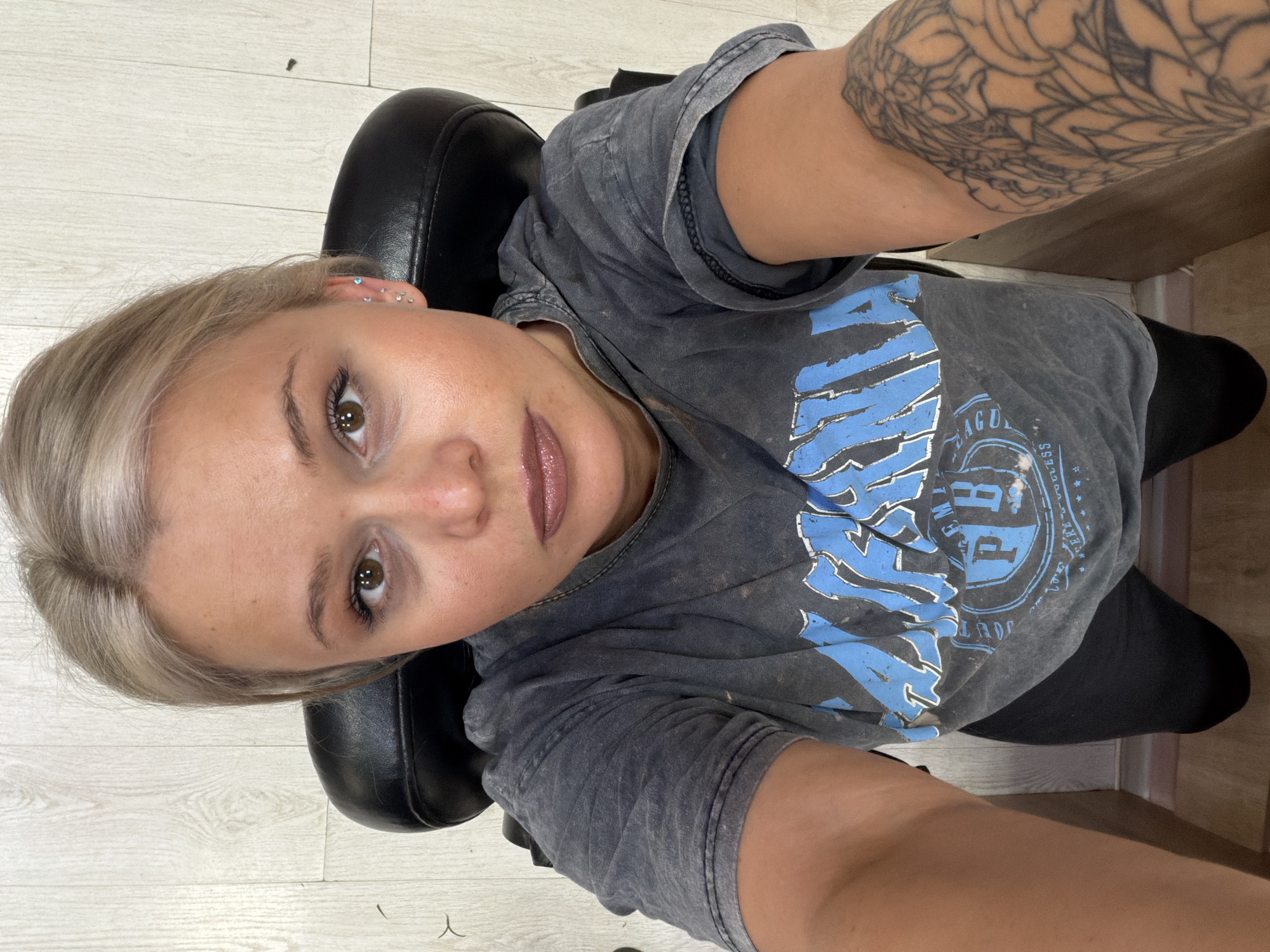 A Caucasian woman is positioned in a dark chair against a backdrop of light-colored, likely wood-grain flooring. Her hair is light blonde, and her skin tone is fair. She has multiple ear piercings. She is wearing a gray graphic t-shirt. A tattoo is visible on her upper arm. The location in Sarapul, Russia, cannot be determined.