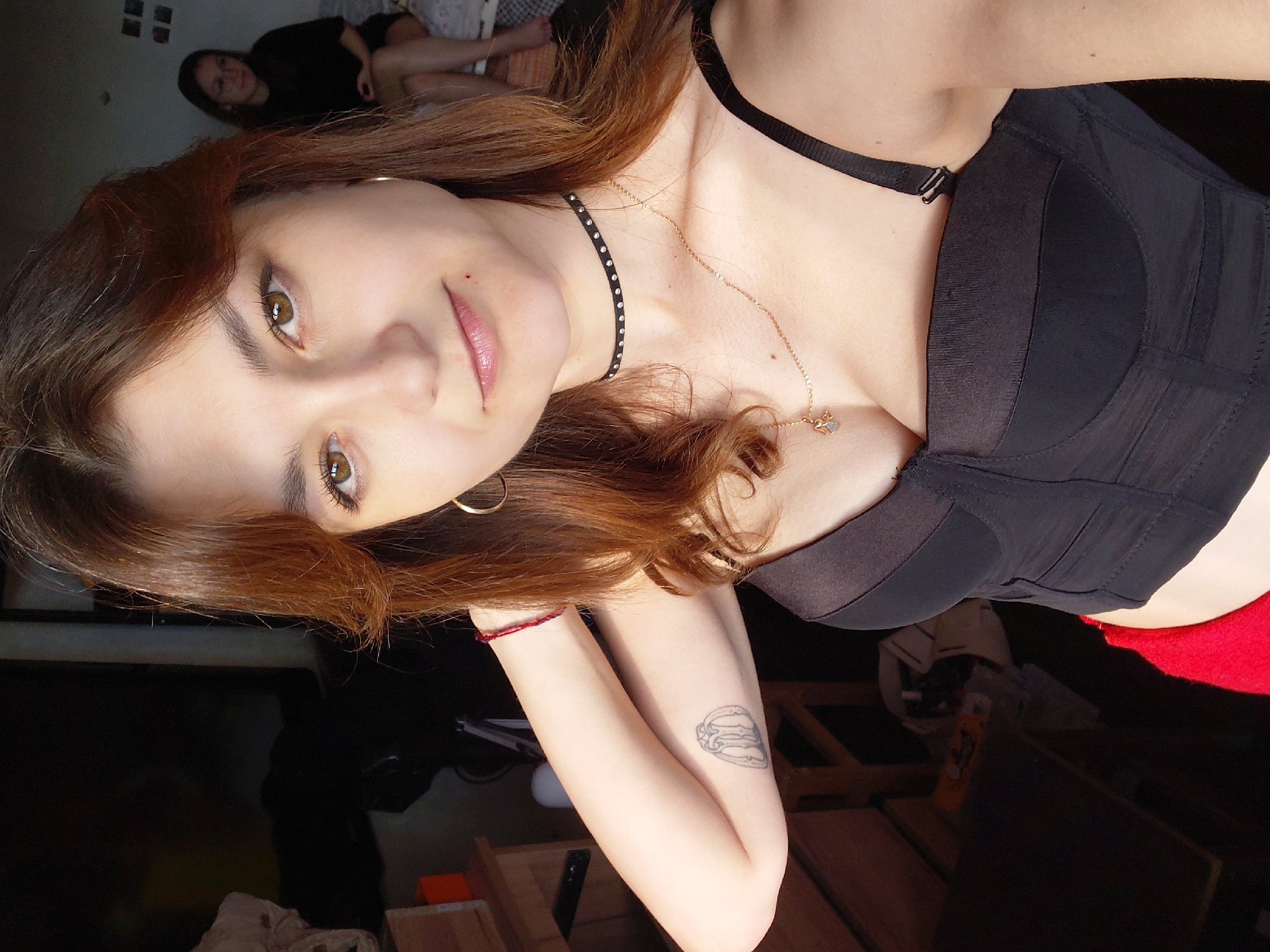 This is a selfie of a young woman with long, auburn hair and light brown eyes. She is wearing a black bra and a delicate necklace. A faint tattoo is visible on her upper arm. In the background, another person is lying on a bed, partially obscured by darkness. The setting appears to be an indoor space, possibly a bedroom, with furniture and clutter visible. The lighting suggests it might be daytime, but the overall mood is intimate and personal, characteristic of a private moment captured by the subject herself. There is no visible text in the image. The location context of Magnitogorsk, Russia, does not provide any specific visual cues within the image itself.