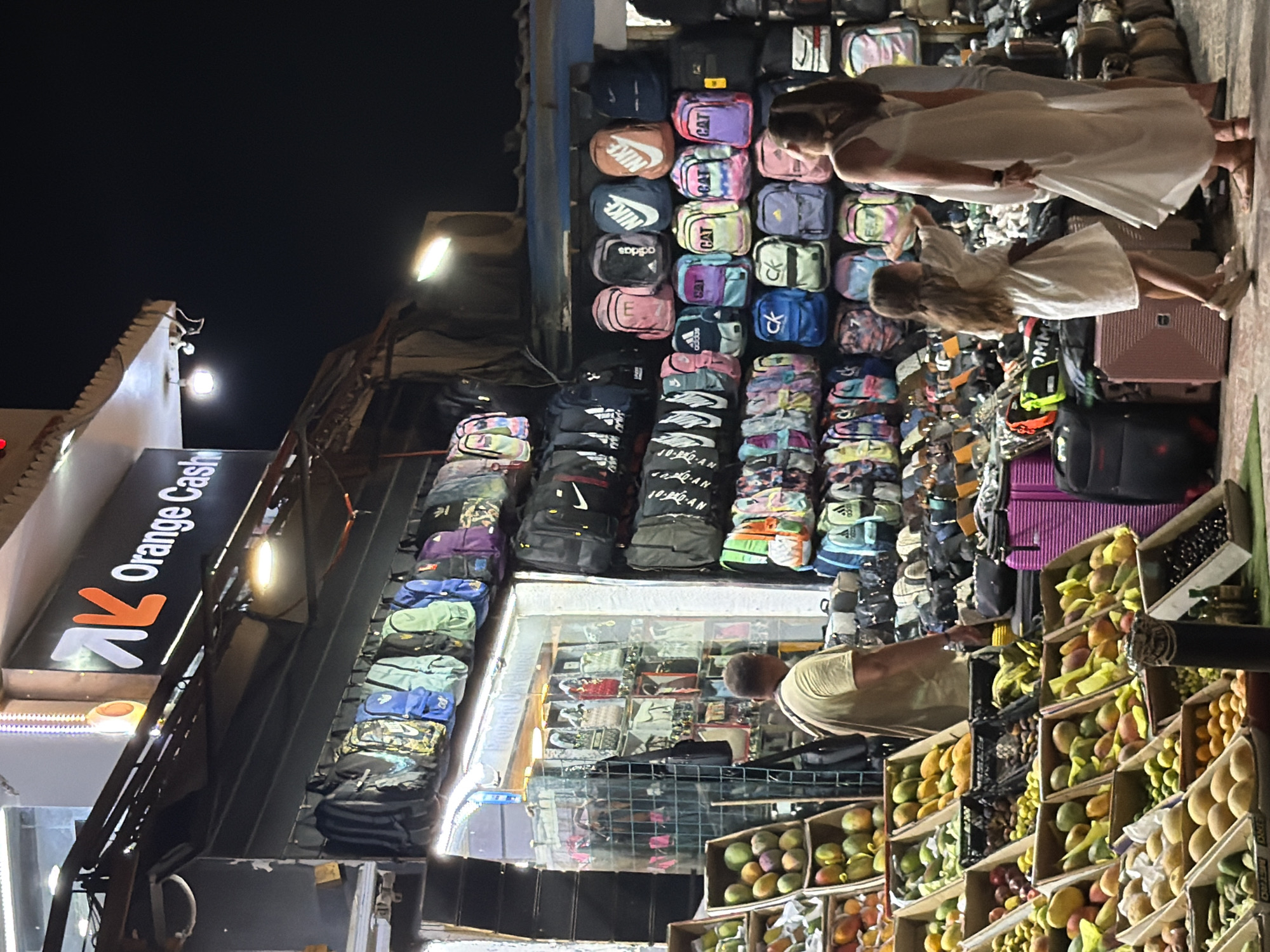 In Sharm El Sheikh, Egypt, an outdoor marketplace at night displays a variety of goods, primarily backpacks and fruit. The scene is illuminated by artificial lighting, casting shadows and highlighting the colorful merchandise.
In the foreground, an assortment of fruits, including mangoes, limes, and possibly oranges, is meticulously arranged in wooden crates. The fruits are of various ripeness and colors, suggesting a bustling fruit stand.
Behind the fruit stand, a man in a tan t-shirt stands facing away from the camera, possibly a vendor or shopper. To his right, a stall is packed with numerous backpacks hanging from a rack and stacked on shelves. The backpacks are diverse in color and design, with visible branding such as Adidas and Nike.
Further to the right, a young girl in a white dress and two women in light-colored clothing are browsing the merchandise. The girl is reaching out towards something, while the women appear to be engaged in conversation or shopping. Adjacent to them, more backpacks are displayed, along with several large suitcases, one of which is a bright pink.
The background reveals the facade of a shop with a sign that reads "AK Orange Cash". The overall environment suggests a lively commercial area in Sharm El Sheikh, likely a tourist-oriented market.