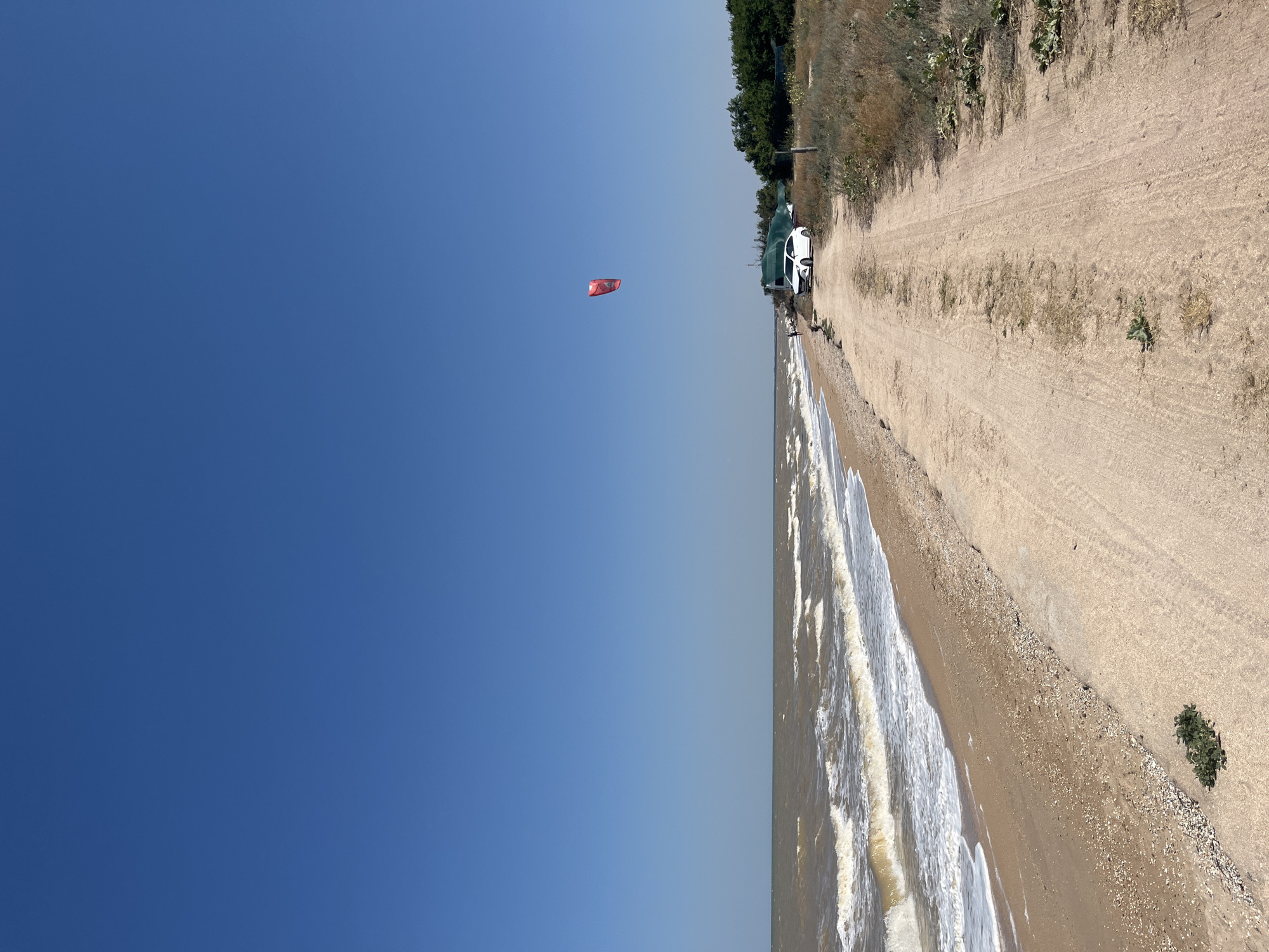 The image depicts a view of the Sea of Azov coastline near Dolzhanskaya, Russia. A sandy beach borders the choppy, brown water. A dirt road runs parallel to the beach, with dry grass and sparse vegetation on its right side. A white car is parked on the side of the road near a structure covered with a green tarp. A bright red kite is visible in the clear blue sky. The scene suggests a daytime environment with strong sunlight, typical of a summer day.