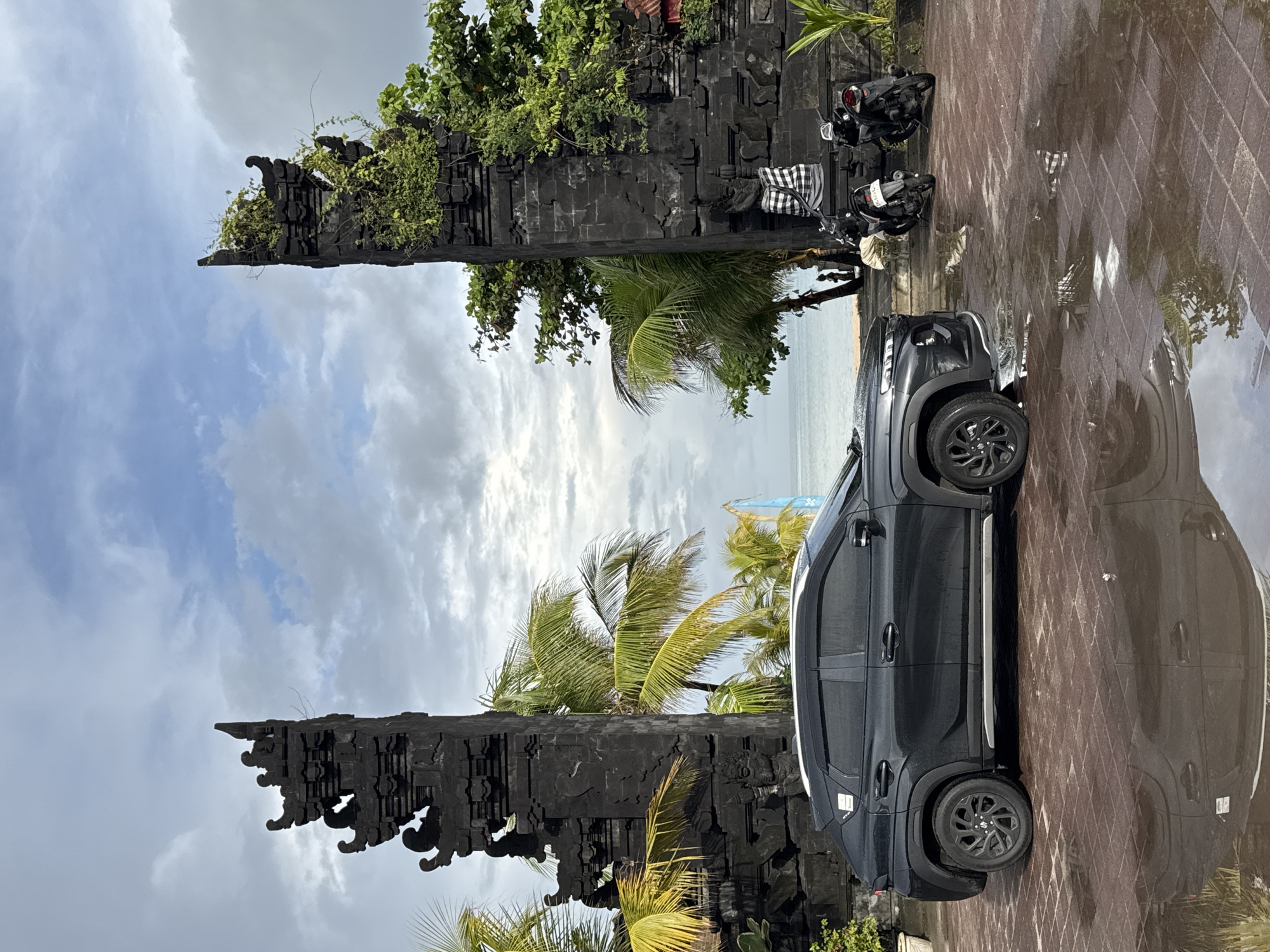 A black SUV is parked on a wet, patterned brick pavement in a coastal setting, likely in Kuta, Indonesia. To the left of the vehicle are two tall, dark, intricately carved stone gates, characteristic of Balinese architecture (Candi Bentar), partially covered with green vines and foliage. Lush palm trees and other tropical vegetation surround these gates and extend into the background.
Beyond the trees and gates, a calm expanse of ocean is visible under an overcast sky with scattered white clouds. The paved ground reflects the car and surrounding elements, indicating it is wet, possibly from recent rain. Two scooters are parked near the gates, one of which has a black and white checkered poleng cloth draped over it. The time of day appears to be daytime, under bright but cloudy conditions. No specific activity is occurring beyond the vehicles being parked. There is no discernible text visible in the image.