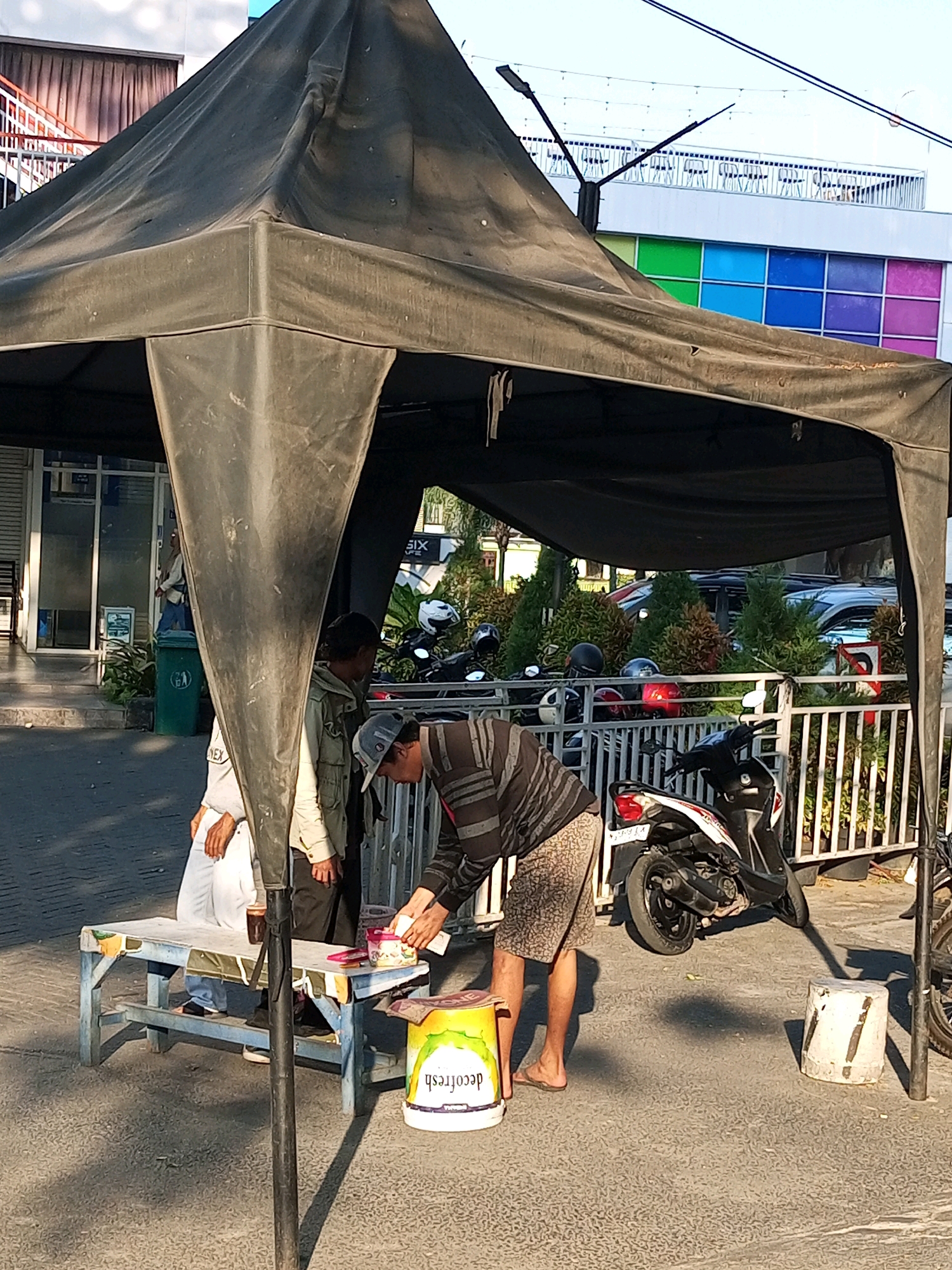 In Batu, Indonesia, an outdoor scene is depicted under a large, dark canopy. Two men are present. One man, wearing a striped long-sleeved shirt, patterned shorts, and a cap, is leaning over a table, appearing to handle items. To his left, another man, wearing a light-colored jacket and dark pants, stands beside the table. A low blue wooden bench is positioned in front of them, with a person's legs visible underneath, dressed in white pants and blue jeans.
On the table, there are various objects including what appears to be a cup, some containers, and possibly paper. A yellow bucket with the label "decofresh" is on the ground to the right of the table. Behind the table, a white metal fence encloses a small area with several motorcycles parked. In the background, there are trees, cars, and a building with a facade featuring a grid of colored squares in green, blue, and purple. The ground is paved. The overall lighting suggests it is daytime with shadows cast on the ground.
