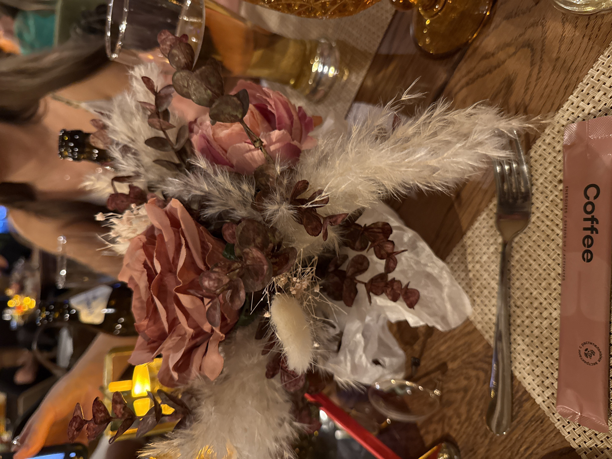 The image shows a table setting, likely in a restaurant. At the center is an arrangement of pink and brown flowers with feathery white accents in a white paper base. Next to the flowers is a fork on a woven placemat. To the right of the fork, on the placemat, is a pink packet labeled "Coffee". A woman and glassware are partially visible in the background. The environment includes a wooden table and other patrons of the restaurant. There is no direct evidence to confirm the location is Peresvet, Russia.