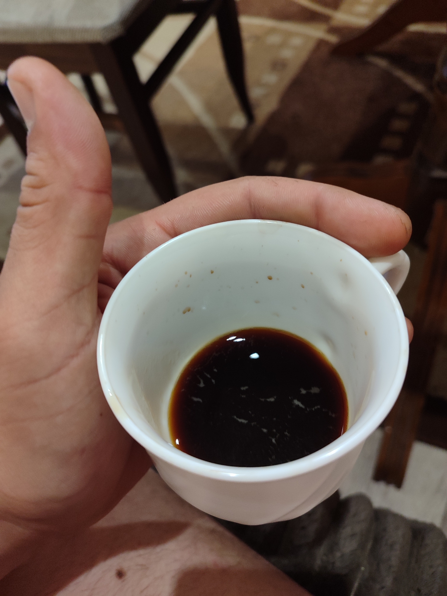 A human hand holds a white cup containing a small amount of dark liquid, likely coffee or espresso. The cup appears used, with residue marks inside. The background is blurred but suggests a room setting with a chair and table visible. No specific details indicate the location being Burgas, Bulgaria.