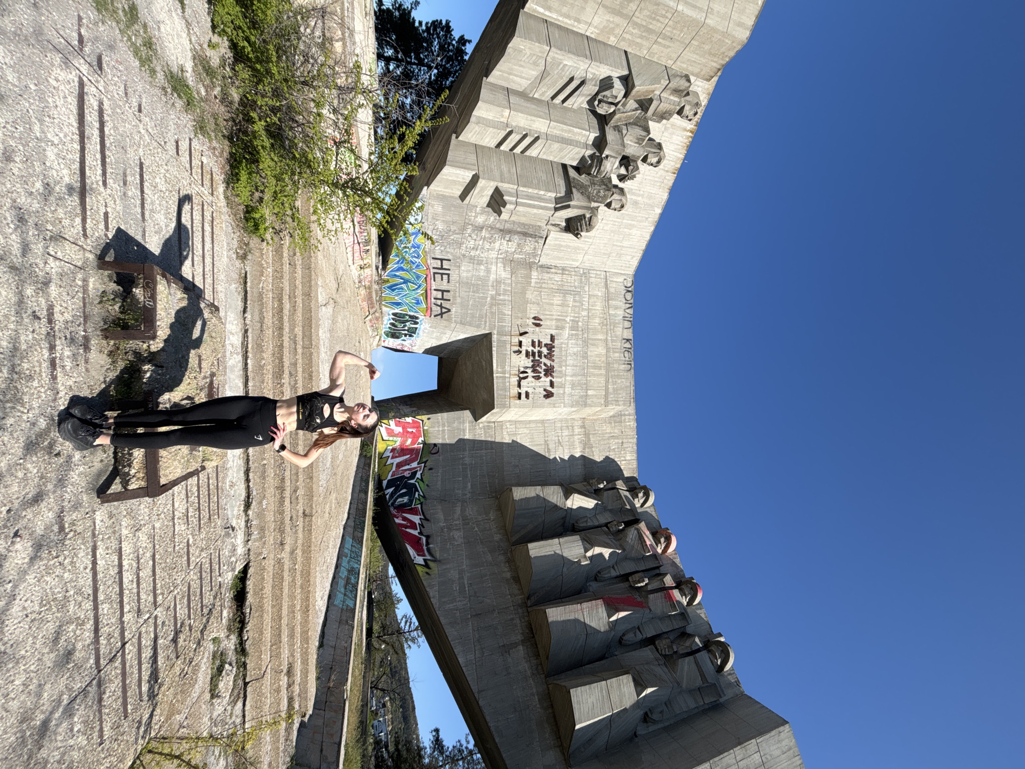 A full-body outdoor shot captures a young woman posing confidently next to a massive brutalist concrete monument under a clear blue sky. The image is oriented vertically, showing the woman standing on a textured concrete or paved surface, which features subtle horizontal grooves, possibly steps. To her left, a simple metal bench casts a long shadow. The woman is dressed in black athletic wear, including a crop top and leggings, and black sneakers. Her arms are bent, hands raised to her head, and her expression is poised.
The monumental concrete structure dominates the right side of the frame, characterized by its sharp angles, geometric forms, and abstract, stylized figures or carvings. It appears weathered and bears various graffiti tags. Prominently visible on one section of the monument are the Cyrillic letters "НЕ НА" (translating to "NO TO") in large black text over a colorful blue and white graffiti pattern. Further to the right, other graffiti includes "COVID KEM" and less legible markings. Scattered green bushes and trees are visible in the background and foreground, providing a natural contrast to the concrete. The bright sunshine indicates it is daytime, casting distinct shadows on the ground and the monument's facets, highlighting its rugged texture. The overall setting suggests a historical site, possibly a memorial, that has been integrated into an urban or natural landscape. Faint text on the bench appears to read "CLAY LO".