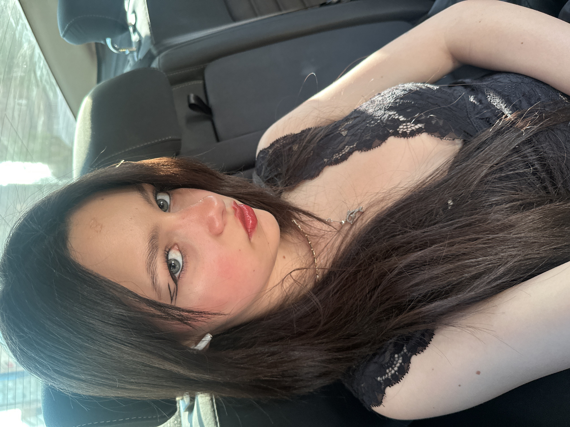 A young woman is reclining on what appears to be the back seat of a vehicle. She is wearing a black lace top and a necklace. The car's interior is black, and there is a window visible in the background. The woman has long dark hair, fair skin, and red lipstick. The location cannot be determined from this image.
