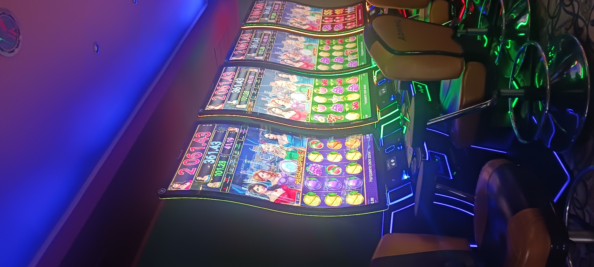 The image shows a row of slot machines in a casino setting, likely in Silistra, Bulgaria. Each machine displays a digital interface with various images, symbols, and numeric readouts indicating potential winnings and betting options. The machines are labeled "Regal Spins 5" and "Admiral." Casino chairs are positioned in front of the machines, illuminated by green and blue neon lights that accentuate the room's ambiance.