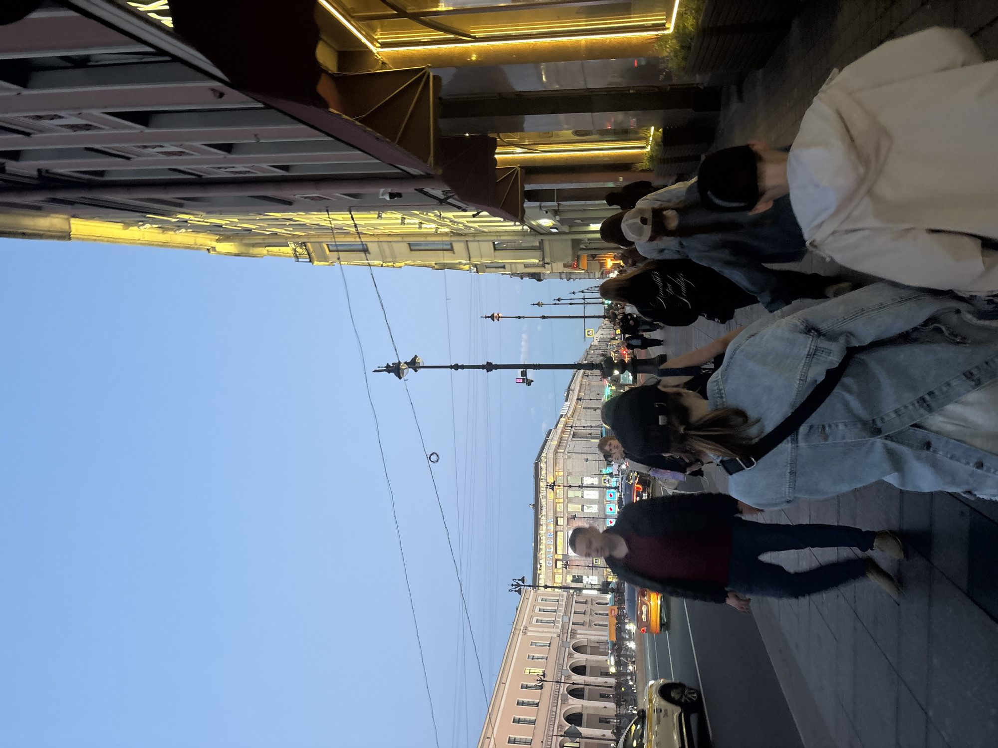 This image depicts a street scene in Saint Petersburg, Russia, likely Nevsky Prospekt, during dusk. A group of pedestrians is walking along the sidewalk. In the foreground, a person wearing a denim jacket is seen from behind, with their hair tied back. To their right, a person in a light-colored hooded jacket is walking away from the camera. Further back, other individuals are walking in the same direction. On the left side of the frame, a street with traffic is visible, including cars and a taxi. The buildings lining the street are illuminated, with some showcasing decorative lighting. The sky is a clear blue, indicating the transition from day to evening. The overall atmosphere suggests a bustling urban environment.