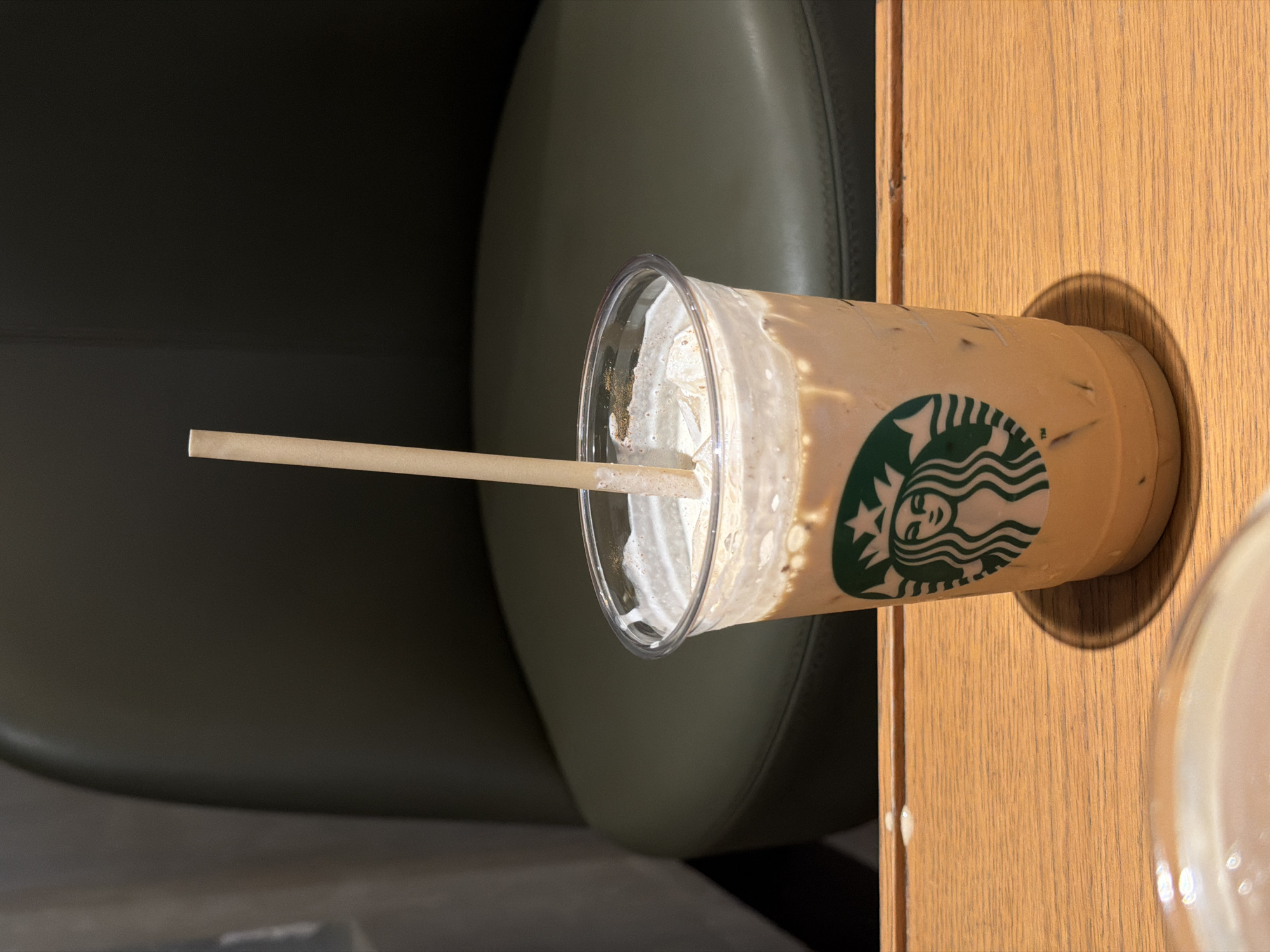 A Starbucks Frappuccino is shown on a wooden table, with a straw sticking out of it. The drink appears to be mostly consumed, with only a small amount of liquid and whipped cream remaining. The Starbucks logo is visible on the side of the cup. In the background, a dark green cushioned chair is partially visible, suggesting the setting is likely a cafe or a similar establishment. The lighting suggests it is daytime. The composition is focused on the drink and its immediate surroundings, with the chair and table providing context for the scene. The overall impression is casual and everyday, perhaps depicting a break or a moment of relaxation.