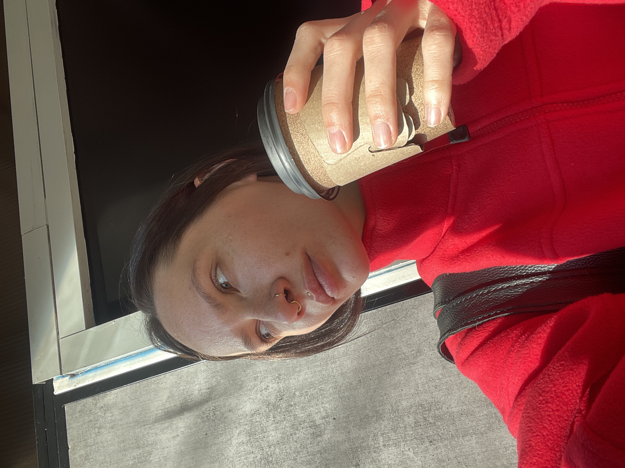 A person wearing a red fleece jacket and a black belt is holding a beverage container. The person has dark hair, fair skin, and a nose ring. The background shows a window frame and an expanse of concrete. This image does not contain the information necessary to determine the location of the photograph. It cannot be determined that this photograph was taken in Izhevsk, Russia.