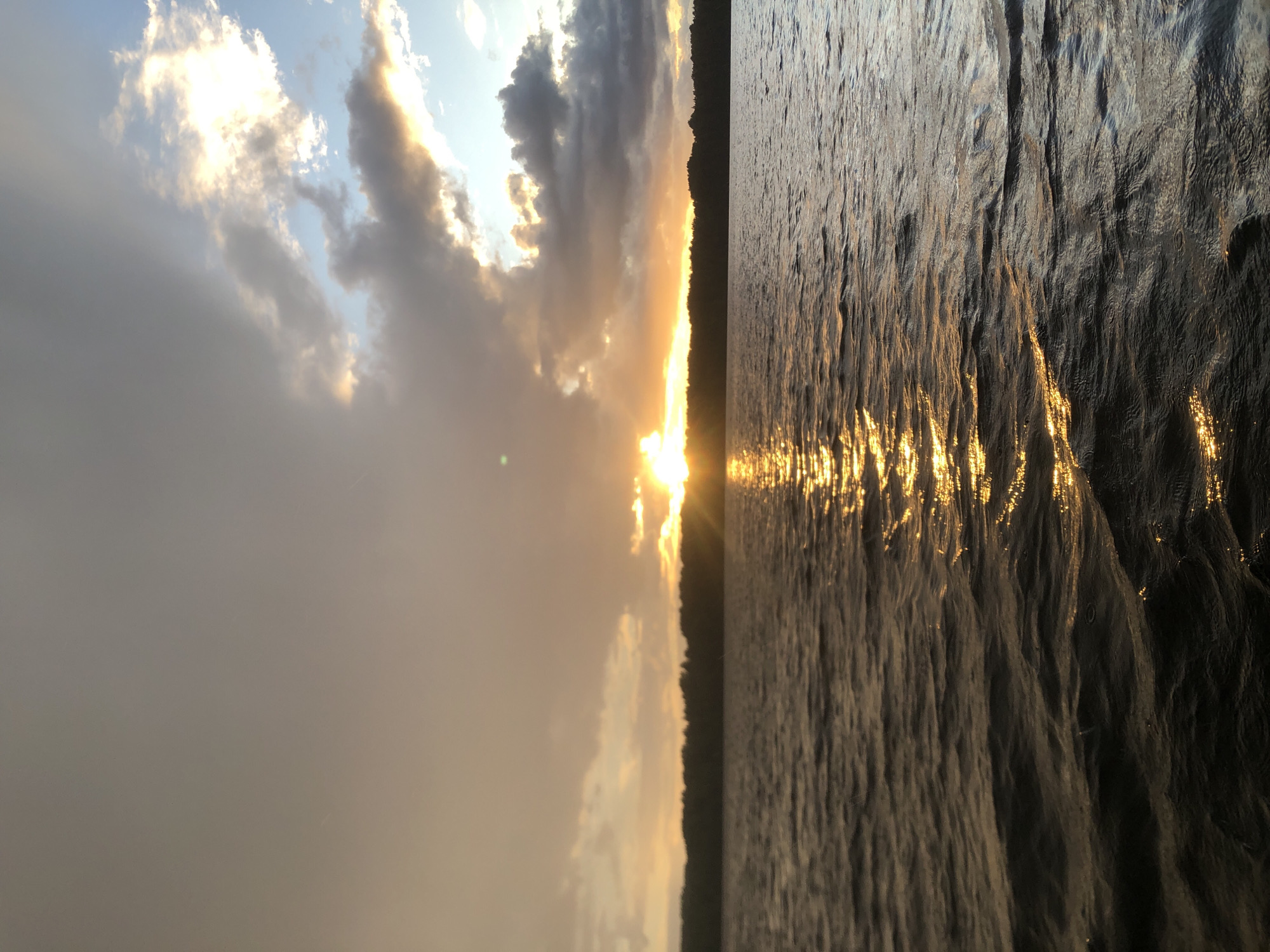 The image depicts a serene sunset over a body of water, likely a river, with a silhouette of trees or a shoreline visible against the horizon. The sun is low in the sky, casting a warm, golden glow that reflects brilliantly on the rippling surface of the water. The sky is a mix of soft blues and grays, with scattered clouds tinged by the sunset's light, creating a dramatic and picturesque scene. The water appears somewhat choppy, with distinct wave patterns catching the light. The overall mood is tranquil and reflective, evoking a sense of peace and natural beauty. The time of day is clearly sunset. There is no visible text in the image. The location is suggested to be Svirstroy, Russia.