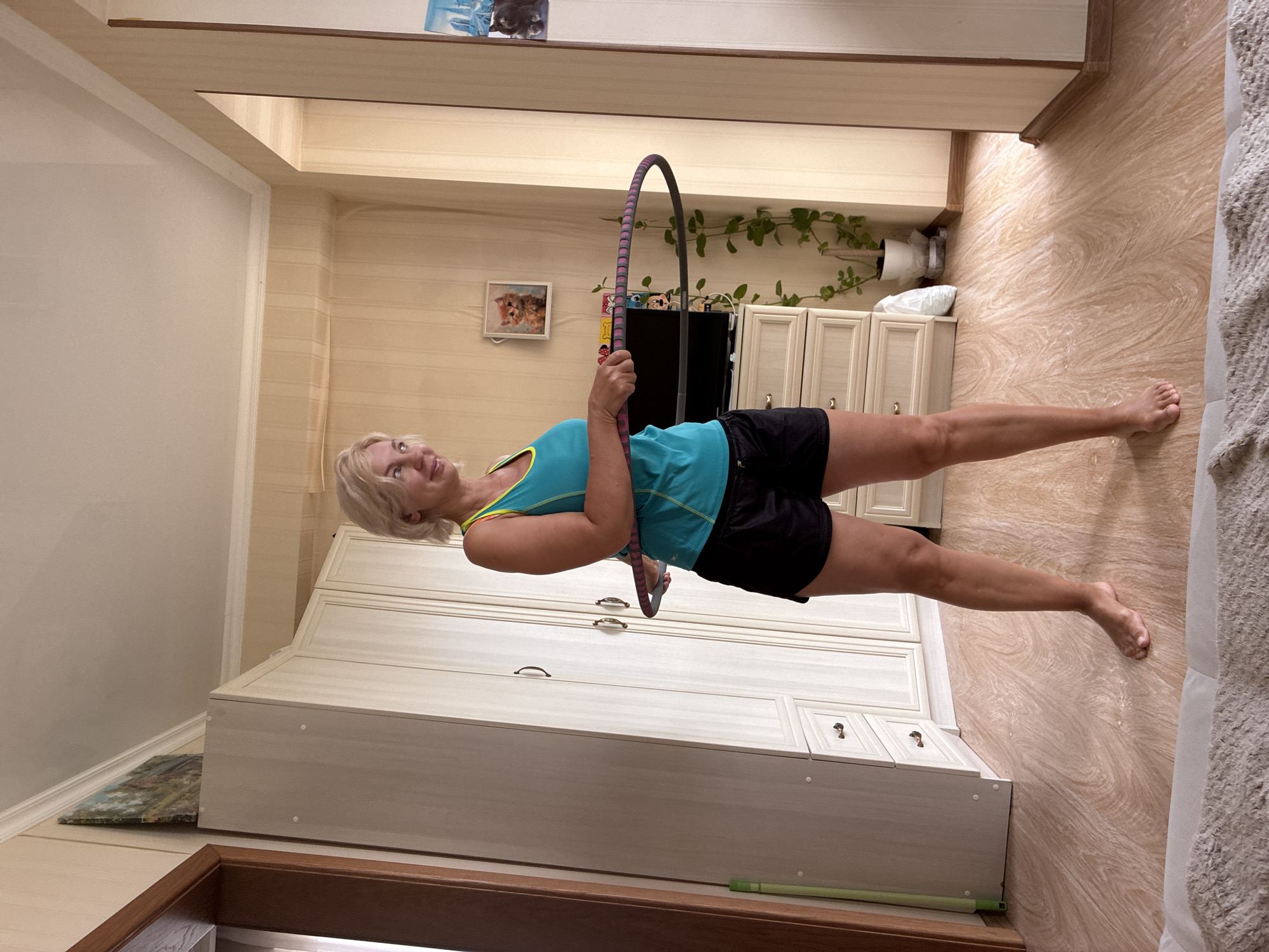 A blonde woman in a turquoise and yellow tank top and black shorts is holding a hula hoop around her waist, appearing to be mid-hula hooping motion. She is standing barefoot on a light-colored wooden floor in a room with cream-colored walls and a light beige patterned wallpaper. To her left is a large, light-colored wardrobe with multiple doors and drawers. To her right, there is a chest of drawers with a television on top, and a potted plant. A framed picture of a cat is hanging on the wall behind the television. The scene appears to be an indoor residential setting, likely an apartment, and there is no visible indication that this location is in the city of Sochi, Russia.