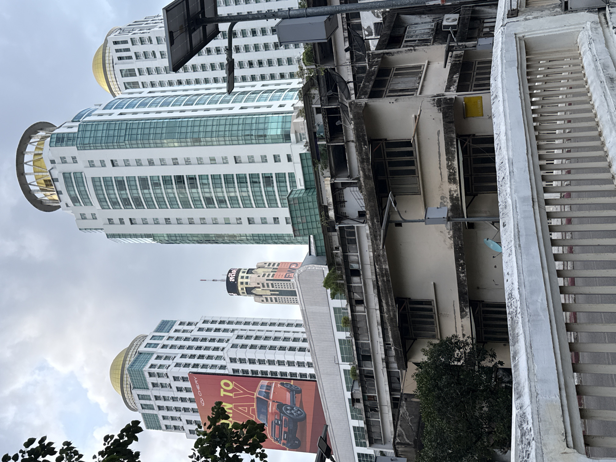 This is a photograph taken in Bangkok, Thailand, focusing on a cluster of buildings under a cloudy sky. The foreground features a white concrete barrier with metal railings, suggesting a raised vantage point, possibly a bridge or overpass. Beyond this, a row of older, dilapidated buildings with beige exteriors and multiple windows are visible. These structures appear weathered, with some windows boarded up or broken. Satellite dishes are mounted on the rooftops of these older buildings.
Behind the older buildings, a series of modern high-rise towers dominate the background. The most prominent are two tall, white skyscrapers topped with golden domes. To their right is another large white skyscraper with green-tinted windows. To the left of the golden-domed buildings, a large billboard displays an advertisement for a red SUV, with the brand "CHERY" and text that appears to read "BORN TO PLAY". Further back and to the right, a uniquely shaped building with an illuminated sign in Thai script is visible, along with the letters "FWD". The overall scene depicts a contrast between older urban decay and modern development in Bangkok.