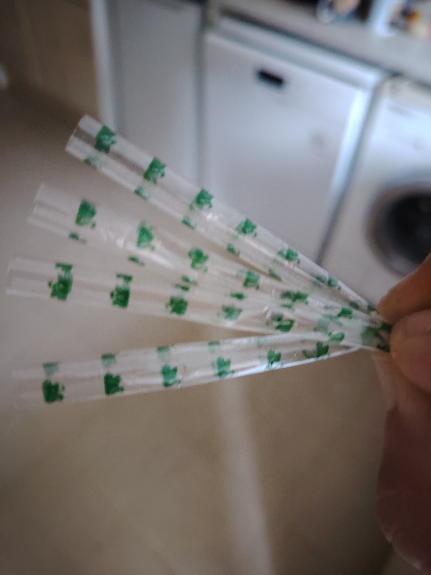 A close-up, slightly blurry, eye-level shot captures a person's hand holding several clear plastic tubes adorned with small green patterns. These tubes are fanned out, with the hand's thumb and forefinger gripping the bottom right edges of the two lowest tubes. The tubes appear to be empty and are arranged in a fan-like formation, with their tips pointing towards the left. In the background, out of focus, are glimpses of kitchen appliances, including what appears to be a washing machine and a refrigerator. The flooring visible is a muted, neutral tone. The scene is indoors, suggesting a domestic setting. There is no specific identifiable location within Huntingdonshire, United Kingdom that can be discerned from the visual evidence.