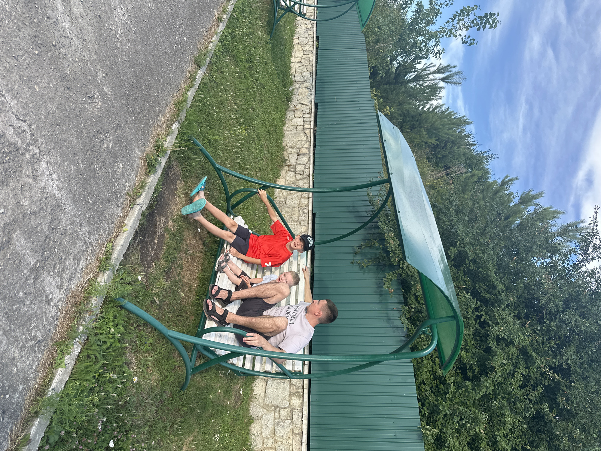 A father and his two sons are sitting on a green metal swing set with a green canopy. The father is wearing a grey t-shirt and black shorts, and the older son is wearing a red t-shirt and black shorts with a black baseball cap. The younger son is wearing a light blue t-shirt and grey shorts. The swing set is on a grassy area with a stone wall and a green metal fence behind it. Trees are visible in the background. The scene appears to be outdoors, likely in a park or recreational area, in Даниловка (алтайский), Russia. The ground in front of the swing set is paved with asphalt.