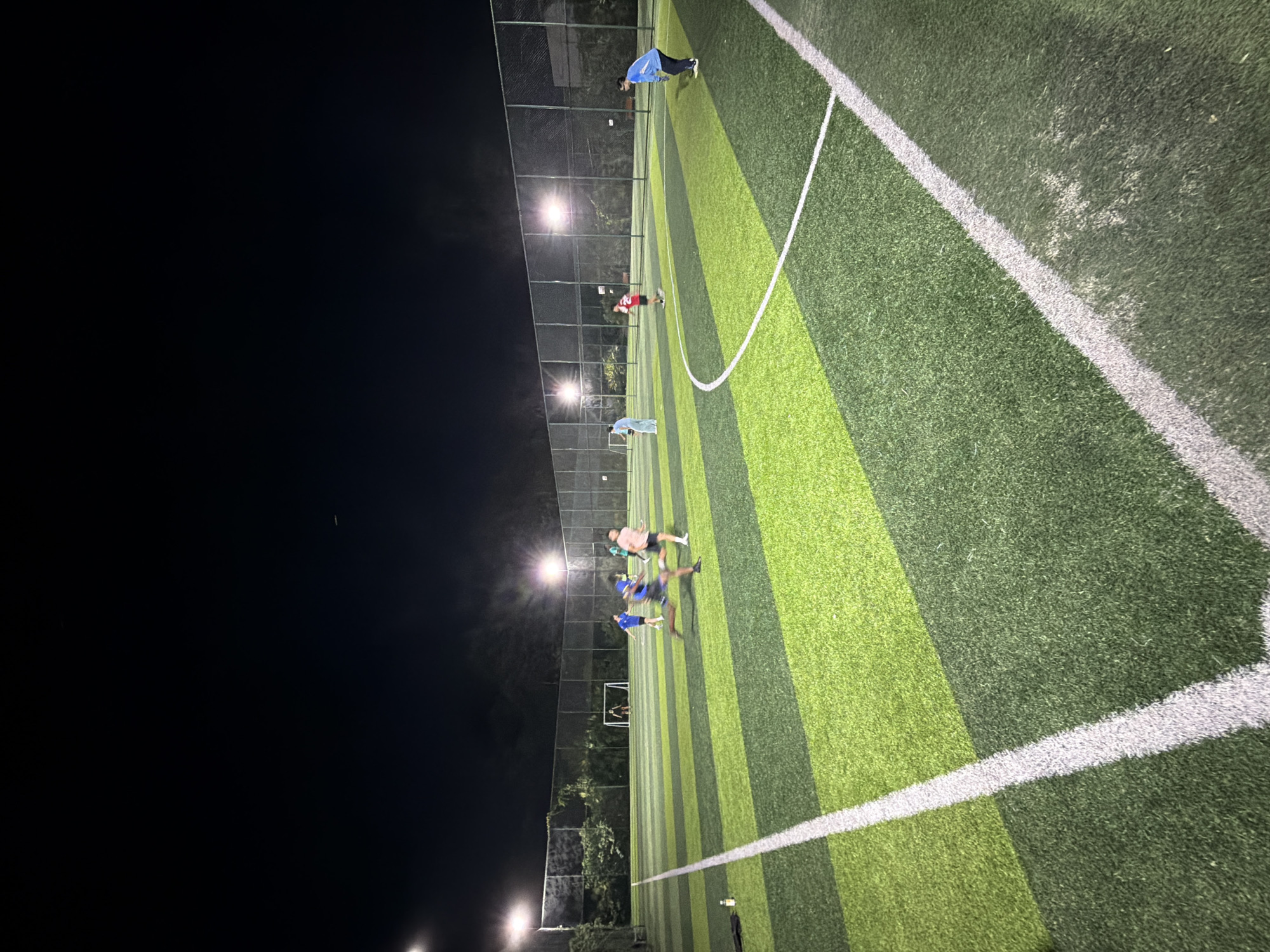 An outdoor, artificial turf soccer field with alternating strips of dark and light green grass is illuminated by bright stadium lights under a dark night sky. A group of six people are engaged in a soccer game. In the foreground, a person in a light blue shirt and dark pants runs towards the right side of the frame. To their left, a person in a blue jersey and dark shorts is in motion, seemingly kicking a ball. Further back, a person in a red shirt and dark shorts is positioned towards the center of the field. On the left side of the field, near the goal, two individuals in blue shirts and dark shorts are near the net. Another person, wearing a light blue shirt and dark pants, stands near the goal on the right side of the field. The field is bordered by a fence and trees can be seen in the background. The image was captured in Jiangjin, China.
