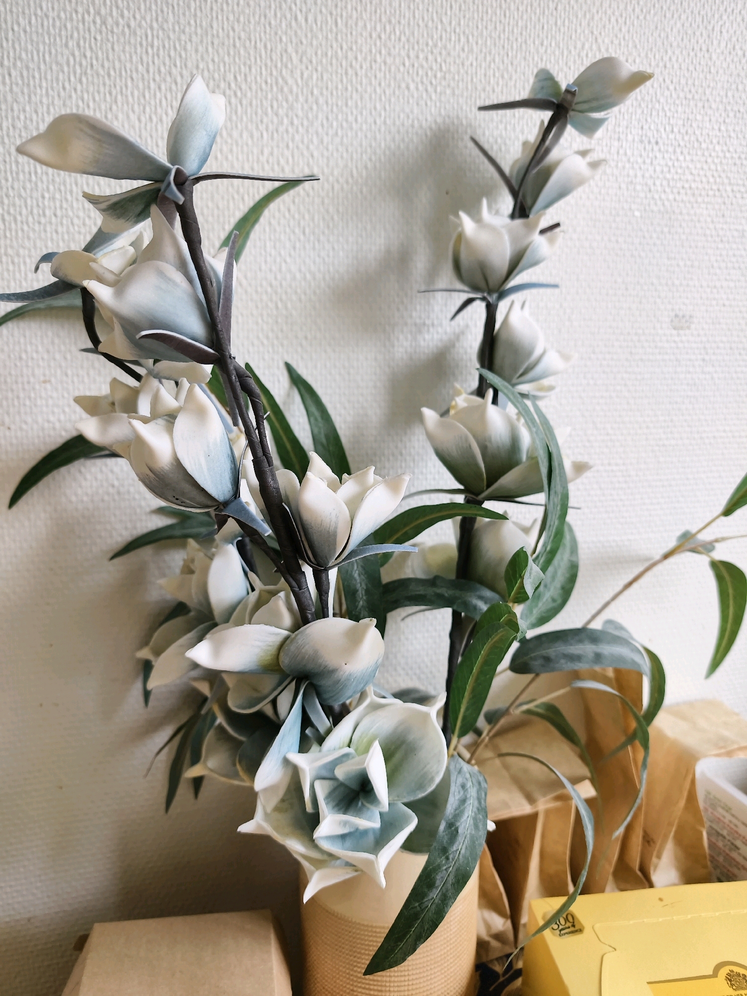 A floral arrangement of white and pale blue flowers with dark green leaves is displayed in a woven vase. The arrangement is placed on a surface with a light-colored, textured wall in the background. To the right of the vase, there are several brown paper bags and a yellow box. The scene appears to be indoors, possibly in a domestic setting or a shop. There are no people visible in the image. This image is not specific to Linköping, Sweden, or any other geographical location.
