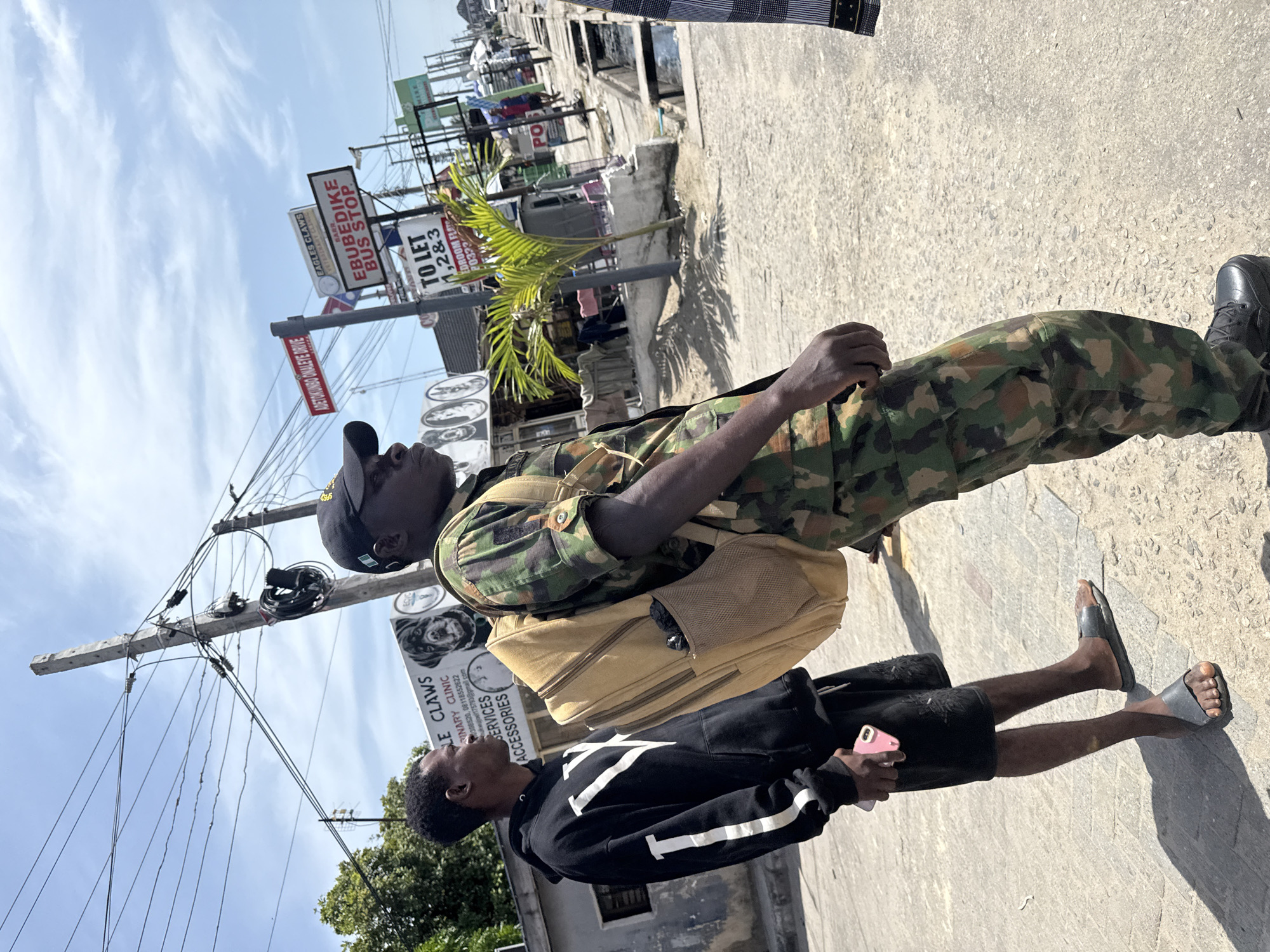This bright, sunlit image captures a street scene in Ado, Nigeria, featuring two men and a backdrop of commercial establishments and utility infrastructure. The man on the right is dressed in a camouflage uniform, complete with a black cap, black boots, and a tan backpack, suggesting he is a soldier or security personnel. He appears to be looking upwards or to his left. The second man, positioned to the left, is casually dressed in a black hoodie with white markings, black shorts, and dark sandals, holding a pink mobile phone in his right hand.
The background reveals a bustling urban environment with numerous overhead electrical wires crisscrossing the sky, supported by a utility pole. Various commercial signs are visible, providing contextual information about the location. Notable signs include "EBUBEDIKE BUS STOP," a "TO LET" sign advertising "1, 2 & 3 BEDROOM FLATS," and a sign for "EAGLES CLAWS VETERINARY CLINIC" which includes phone numbers (08035085220, 08118552622) and an email (175783@gmail.com). Another sign identifies the street as "ADETOKUNBO ONALEYE DRIVE." A small palm tree adds a touch of greenery to the roadside. The clear sky with wispy clouds and prominent shadows indicates a sunny day.