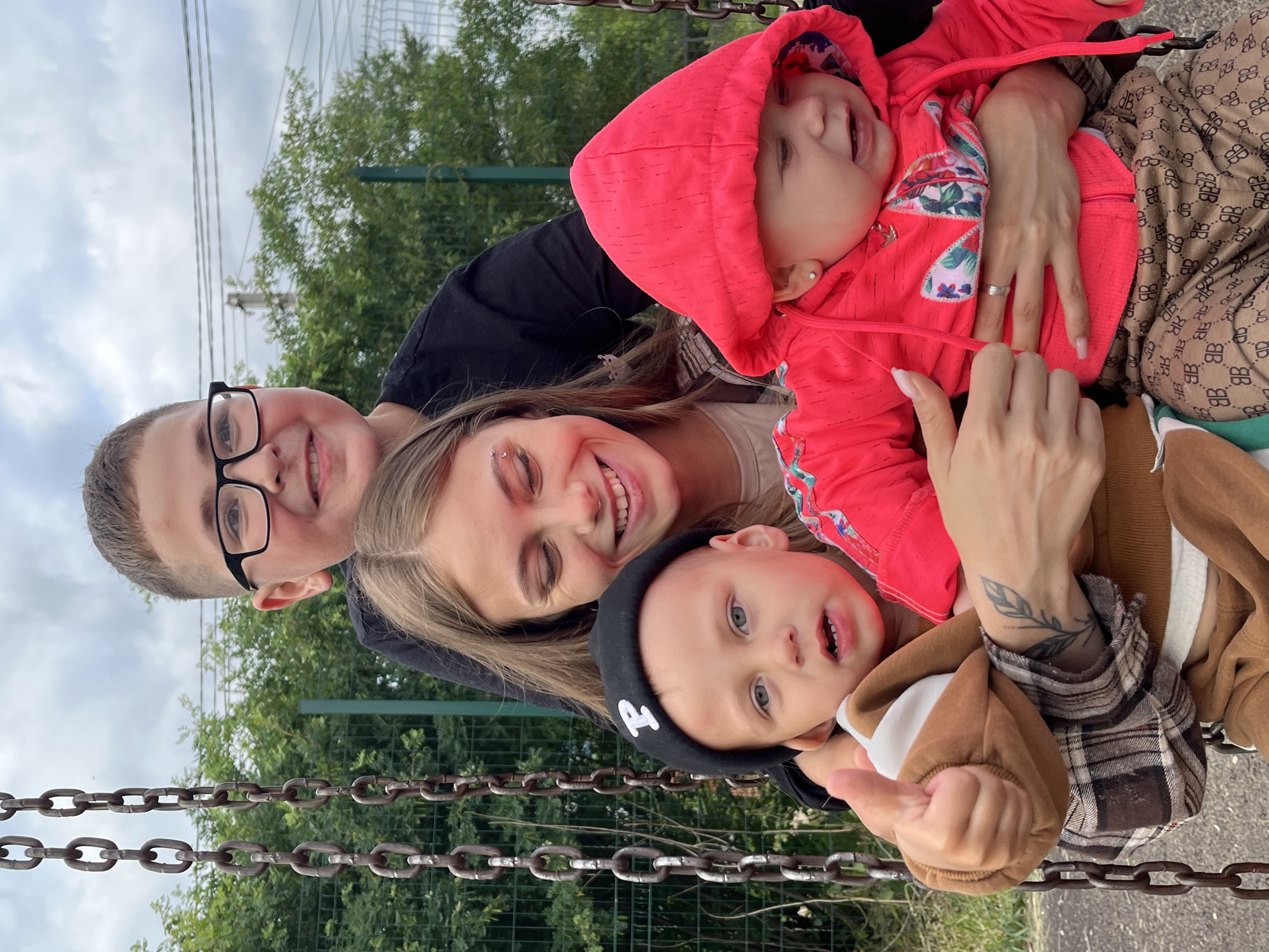 A group of four children, one young woman, and one boy are on a swing set. The young woman is in the middle, holding a baby on her lap. To her left is a young boy wearing a black beanie with a white "P" on it. Behind the young woman and to her right is a toddler in a pink hoodie. Behind the young woman and to her left is a boy wearing glasses and a black shirt. The swing set chains are visible on the left side of the image. The background consists of trees and a fence. The location is the city of Fokino, Russia.