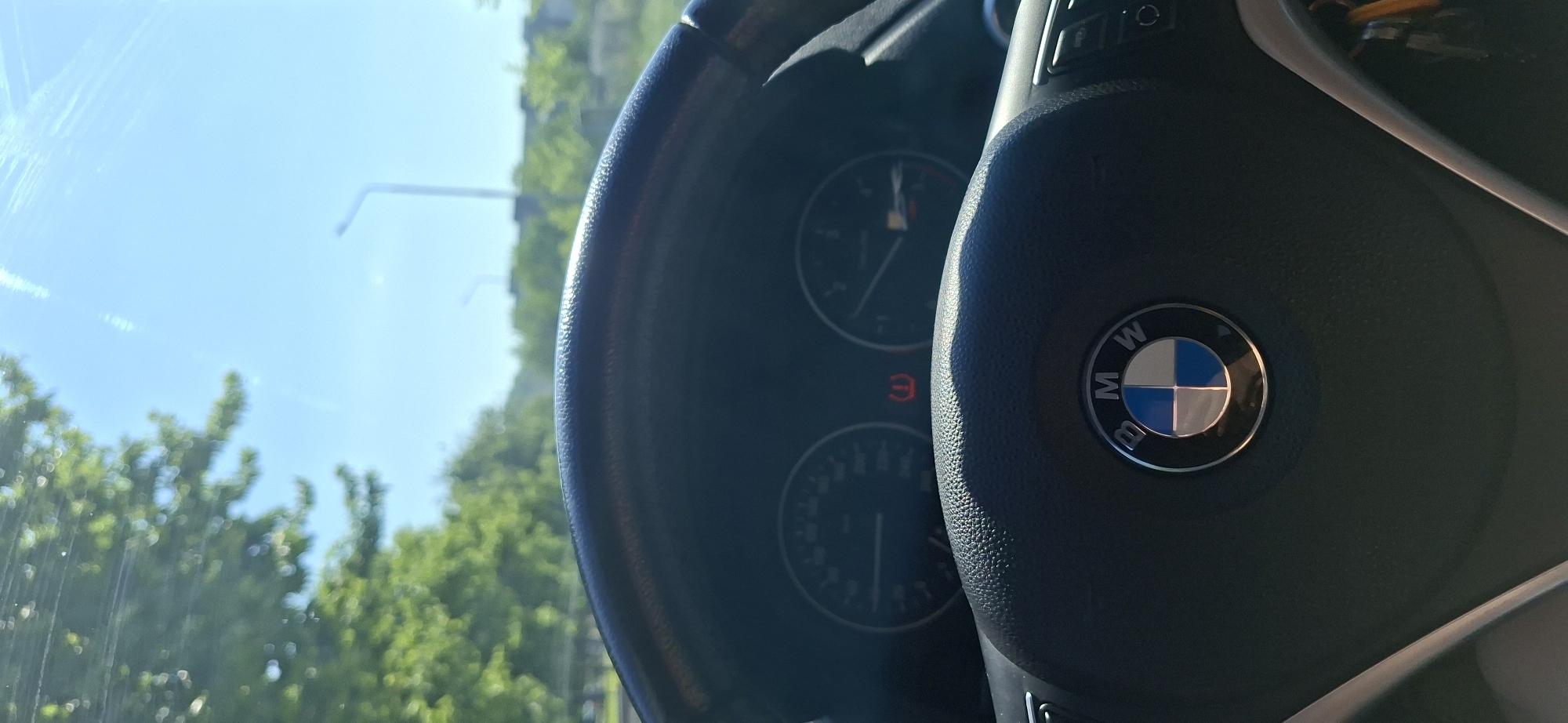 The image shows the interior of a BMW car, focusing on the steering wheel with the BMW logo prominently displayed. The dashboard gauges are partially visible behind the steering wheel. Through the windshield, a view of a street with trees and a street lamp is seen, suggesting an outdoor setting. The scene appears to be from within the car, looking out. Given the prompt's request for a location in Plovdiv, Bulgaria, the scene is presumed to be within the city of Plovdiv, though no specific landmarks are visible to confirm the precise location.