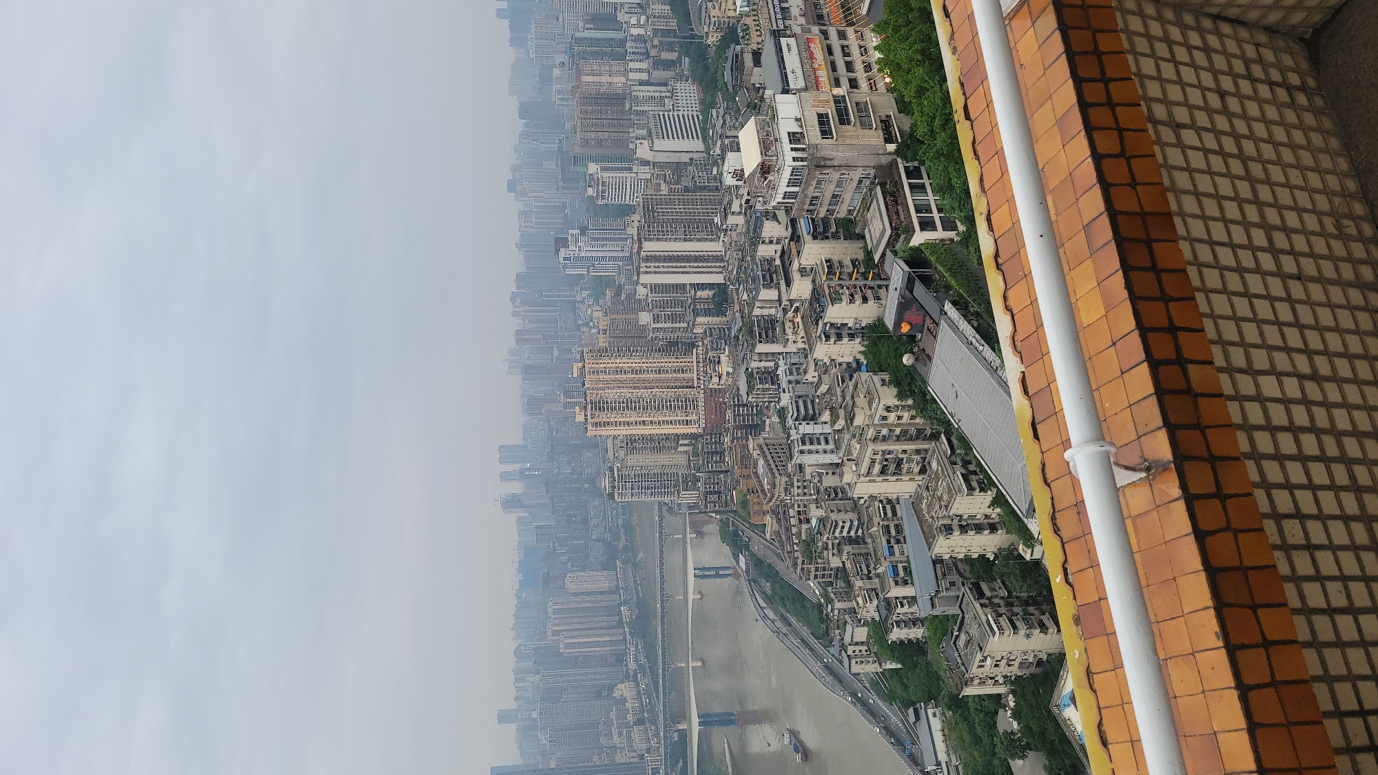The image shows a cityscape viewed from a high vantage point. The city, presumably in Unknown, Unknown, is densely populated with numerous high-rise buildings extending into the distance, partially obscured by haze. A river flows through the city with bridges connecting different parts. The foreground includes a section of a tiled brick wall and a white pipe, likely part of the building from which the photo was taken.