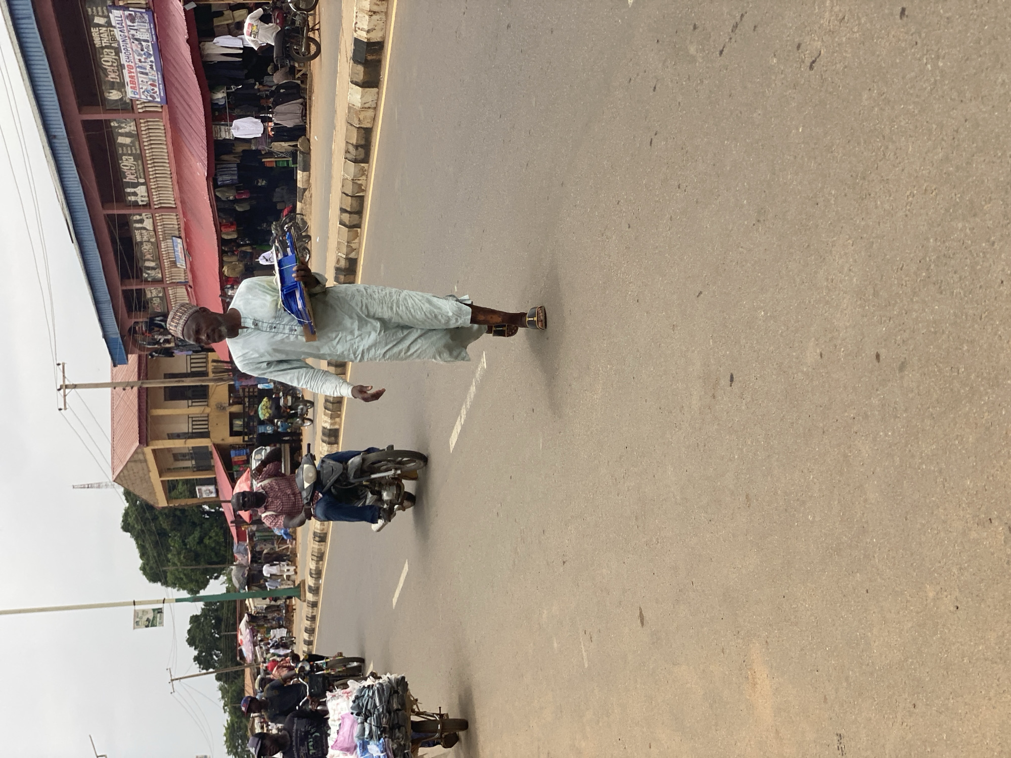 This media file captures a bustling street scene in Kafanchan B, Nigeria, oriented sideways.
In the foreground, a man wearing a light-colored traditional tunic (likely a caftan or similar garment) and a white kufi cap walks confidently in the middle of a paved road. He is holding a blue plastic tray or container in his left hand and appears to be looking towards the camera with a slight smile or friendly expression, extending his right hand slightly. He is wearing sandals.
Behind him, on the road, a person on a motorcycle is visible, moving in the same direction. Further down the road on the left, another motorcycle is heavily laden with what appears to be textile goods or clothing. The road itself is asphalt, with faded white lane markings. A concrete curb separates the road from the unpaved shoulder and storefronts.
On the left side of the street, multiple single and two-story buildings house shops and businesses. Many of these shops display various items, primarily clothing, on racks outside. A prominent blue and white sign on one of the buildings clearly reads "bet9ja" and "BABATU SHOPPING MALL". Other illegible signs are also visible. The buildings feature red corrugated roofs.
The background shows more buildings, green trees, and utility poles with overhead wires, indicating a built-up area. The sky is bright, suggesting daytime, likely late morning or afternoon, with clear weather. The overall scene depicts a typical active market or commercial street in a Nigerian town, characterized by pedestrian traffic, motorcycles, and roadside commerce.