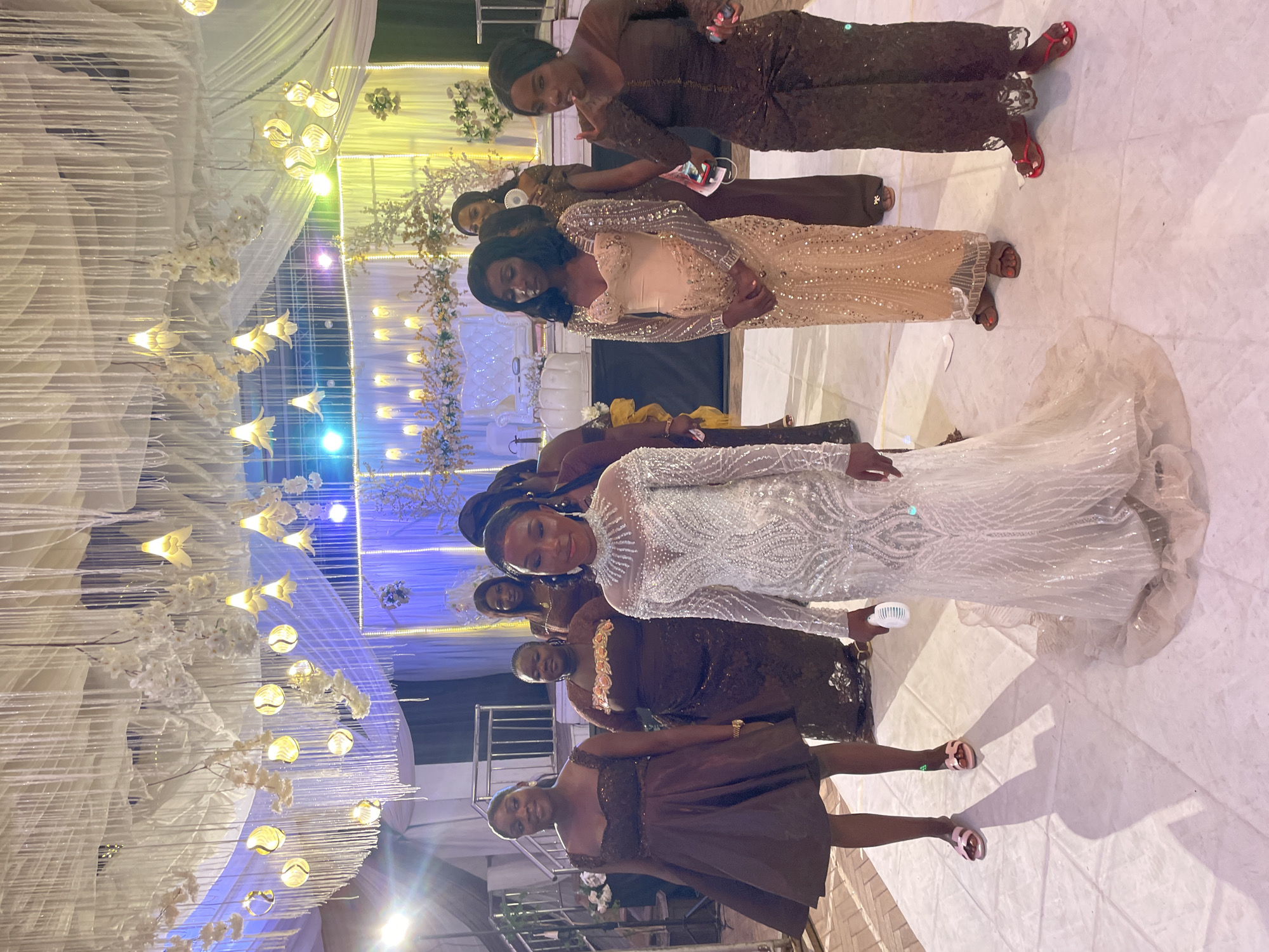 The image shows a group of women indoors, at what appears to be a wedding reception in an unknown city. In the center, a woman in a white wedding dress is surrounded by other women in brown and gold dresses. The women are standing on a light-colored floor. Above them, the ceiling is decorated with white fabric and hanging lights.