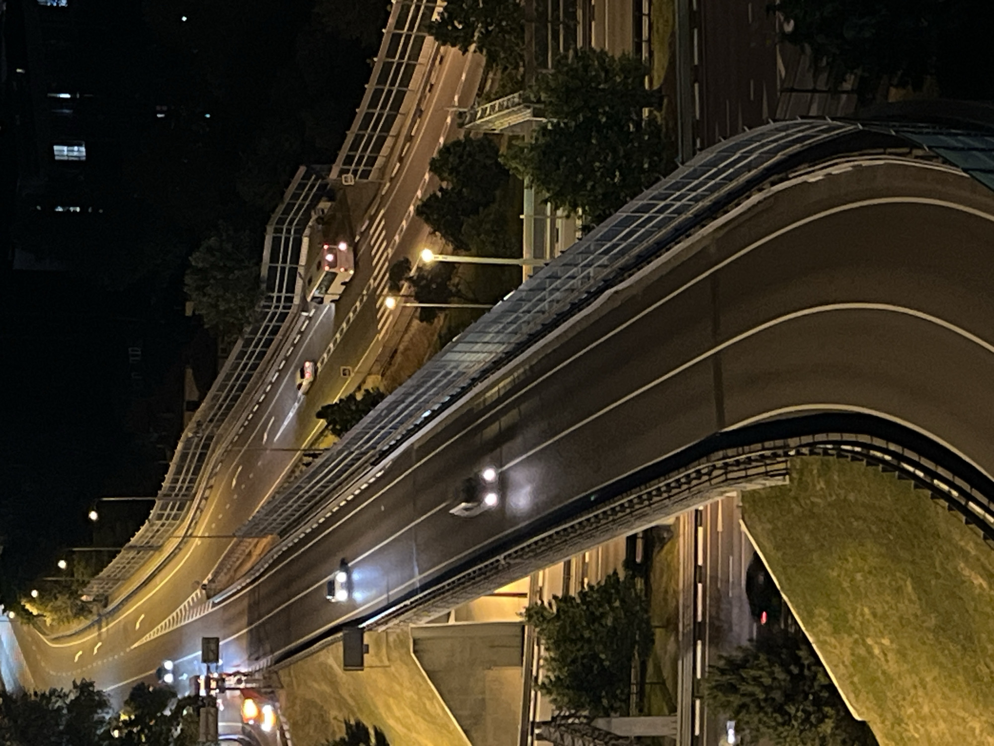 The image shows an elevated highway at night in Sochi, Russia. Cars are driving on the highway, and streetlights illuminate the road. The highway is bordered by a fence and trees. In the background, buildings can be seen.