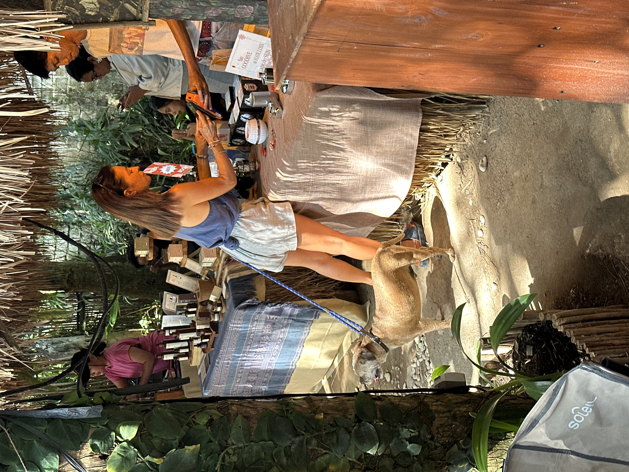 A woman in a blue tank top and striped shorts is standing at a market stall, holding a credit card reader. She is interacting with a man in a patterned shirt who is holding a phone. To her left, a tan dog with a blue leash stands on the dusty ground. Behind them, a stall is set up with various products displayed on tables. In the background, more people are visible, and the market appears to be outdoors with a thatched roof overhead. This scene is taking place at a market in North Kuta, Indonesia.