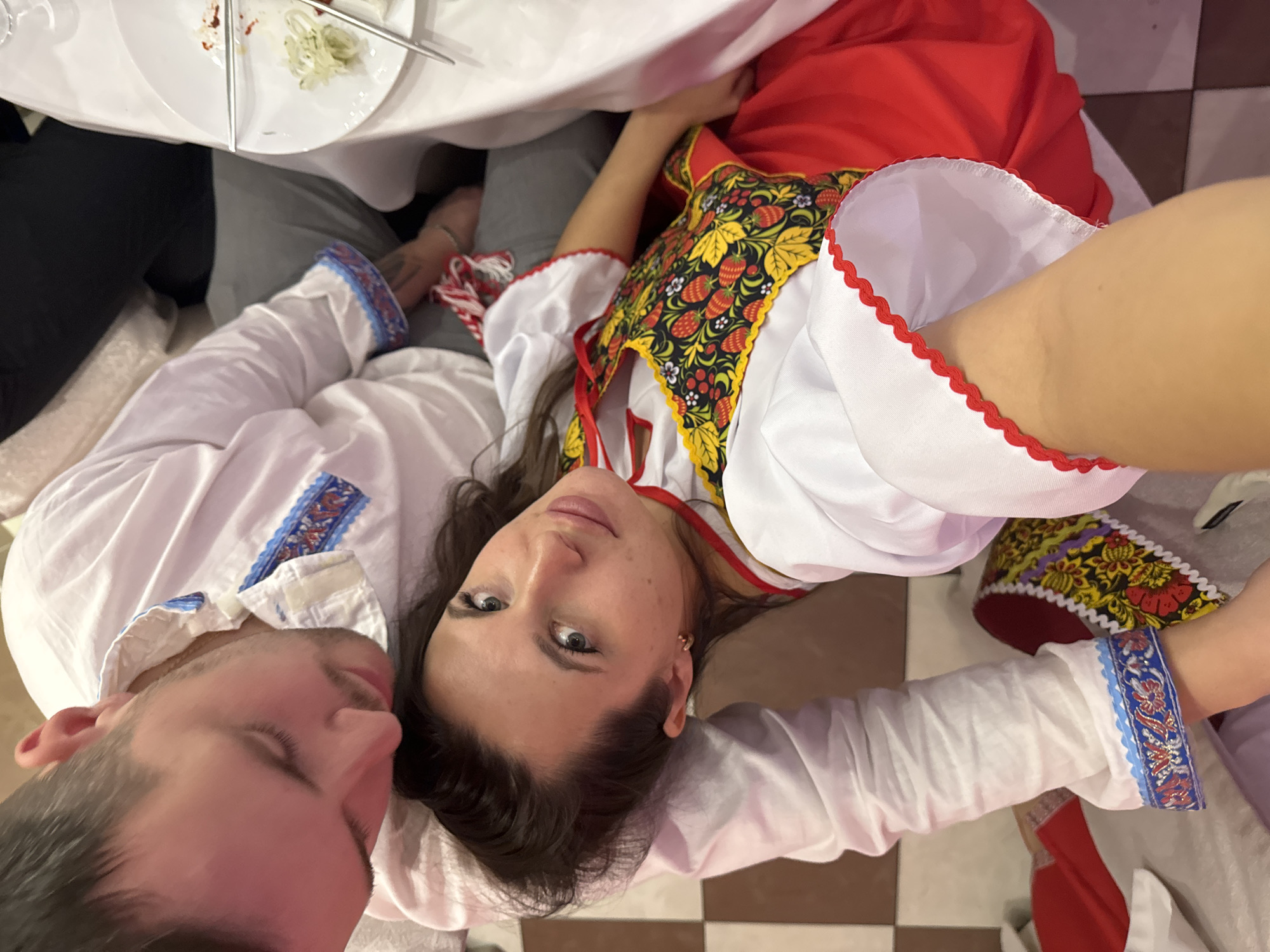 A man and woman are lying down and looking at the camera, likely taking a selfie. The woman is wearing a red and white dress with an intricate floral pattern around the chest. The man is wearing a white shirt with blue trim on the cuffs and collar. A table with dishes is above them. The background appears to be a tiled floor. The city and state are unknown.
