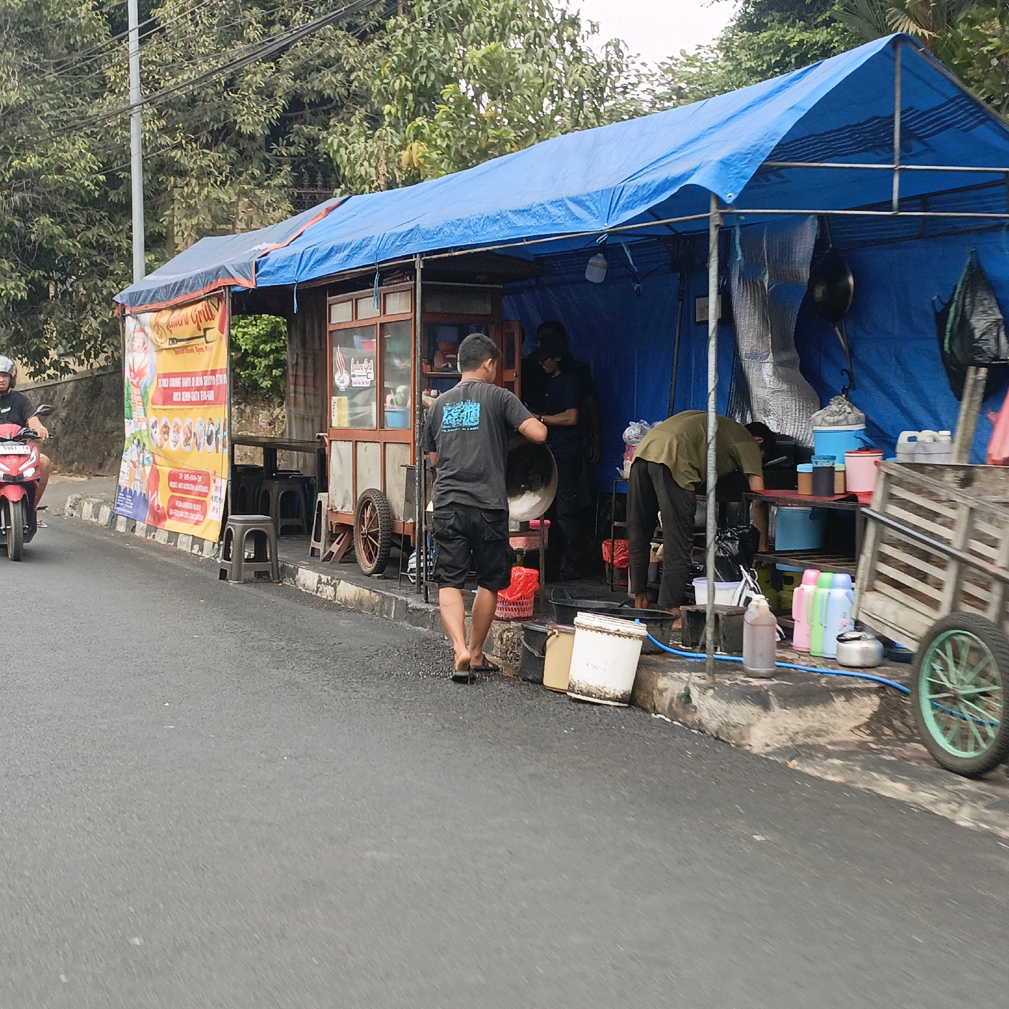 A food stall under a blue tarpaulin is set up on the side of a paved road in Salatiga, Indonesia. The stall appears to be a temporary structure with a wooden counter and a display area behind glass. A large, colorful banner advertising food is displayed on the left side of the stall. Several people are present, some working at the stall and one on a red motorcycle approaching from the left.
In the foreground, the asphalt road is wet, suggesting recent rain or cleaning. The stall itself is equipped with various containers, bottles, and cooking utensils. A man in a black t-shirt and shorts is handling a large metal bowl, possibly preparing food or cleaning. Another person in a dark shirt and cap is visible behind the counter. To the right, a person in a green shirt is bending over, working near shelving units. A wooden cart with a green spoked wheel is parked to the right of the stall. Trees and vegetation are visible in the background, along with a utility pole. The overall scene depicts a bustling street-side food business.