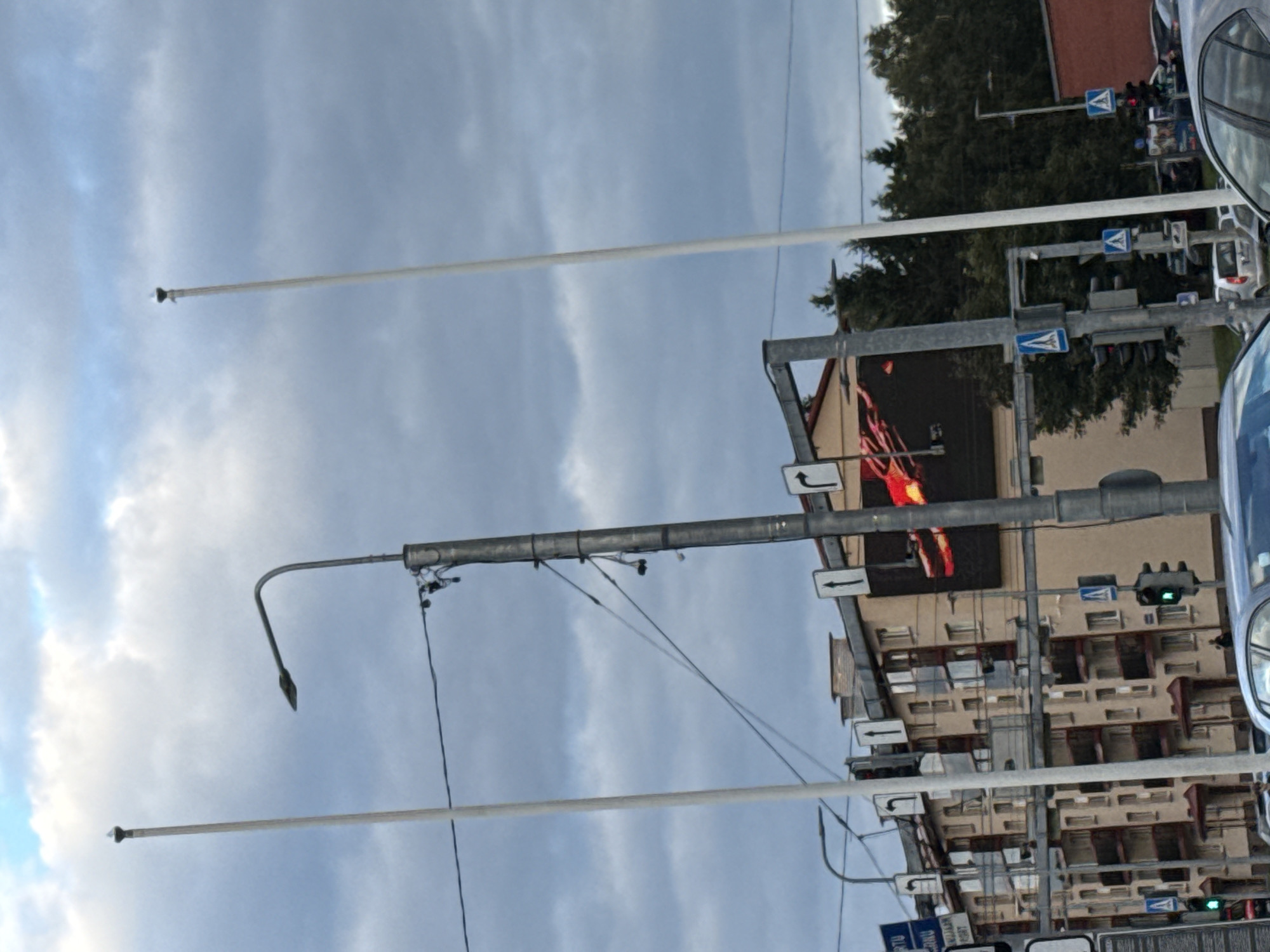 The image captures a street scene in Tallinn, Estonia, under a cloudy sky. In the foreground, a tall, slender white pole stands vertically, while a shorter, gray pole with a streetlamp curves to the left. A traffic light with green illumination is visible. A multi-story building with beige siding and balconies occupies the mid-ground. To the right of the building, a large digital billboard displays a brightly lit, red-toned abstract image. Traffic signs indicating directional arrows are mounted on poles. Several cars are present, including a silver car parked on the street and a dark-colored sedan in the foreground. Trees are visible in the background. The scene appears to be an intersection with pedestrian crossings.
