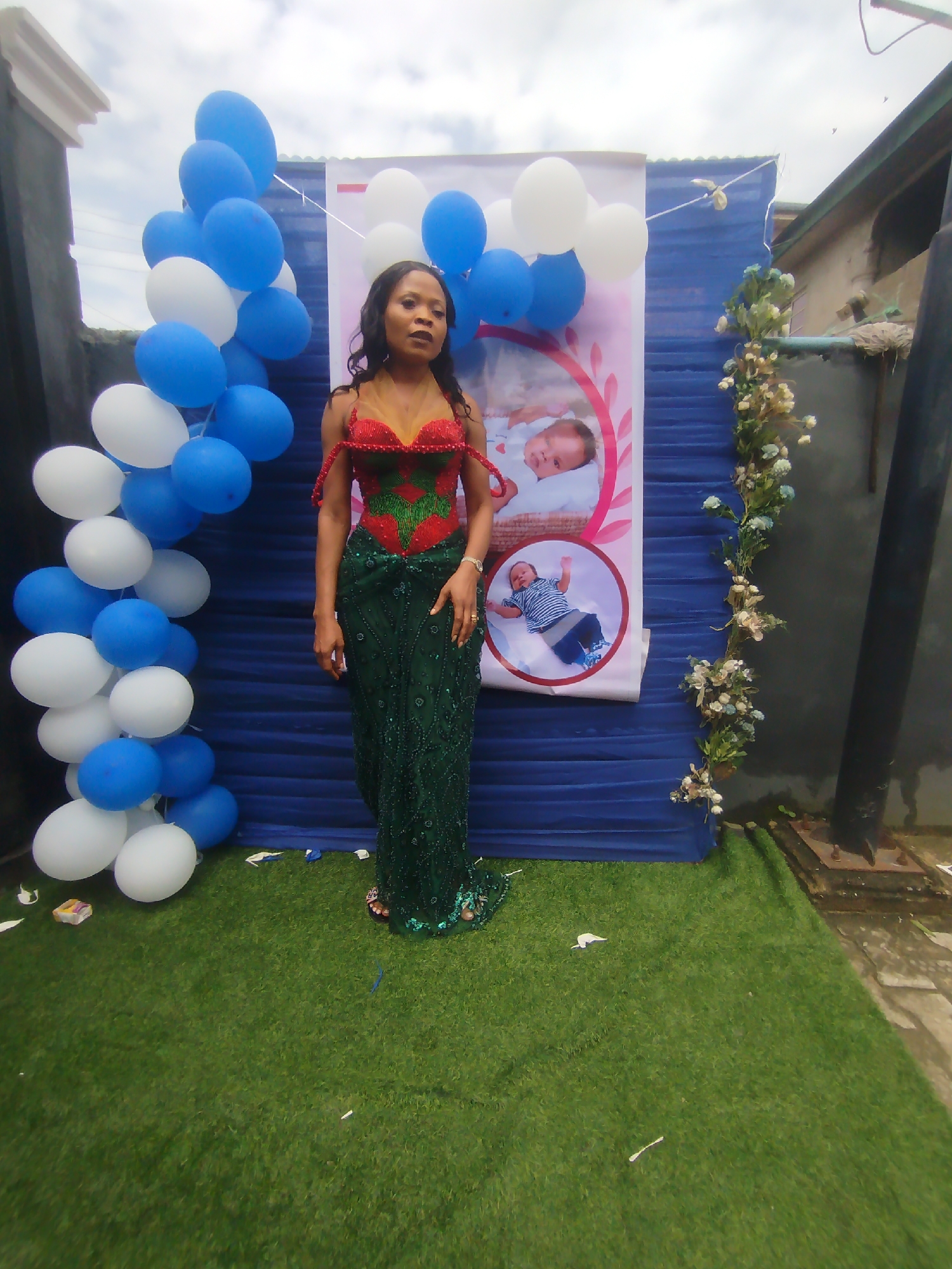 A woman in a green sequined dress with a red and green beaded top stands on artificial grass in front of a blue backdrop. Balloons in blue and white are arranged in a curve to her left. Behind her, a banner displays images of babies. The scene appears to be an outdoor event setup, possibly a naming ceremony or birthday celebration in Yenegoa, Nigeria. A dark pole and a section of a building are visible to the right.