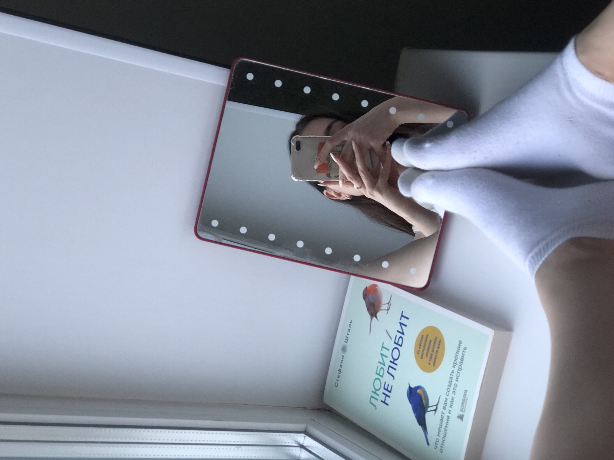 A person, presumably a woman, takes a mirror selfie on a white windowsill in Noviy, Russia, showing white-socked feet and the Russian version of the book "The Child in You Must Find a Home" by Stephanie Stahl, titled "Lubit/Ne Lubit." The selfie is taken with a phone with a white and orange-patterned case. The mirror has a red frame with lights.