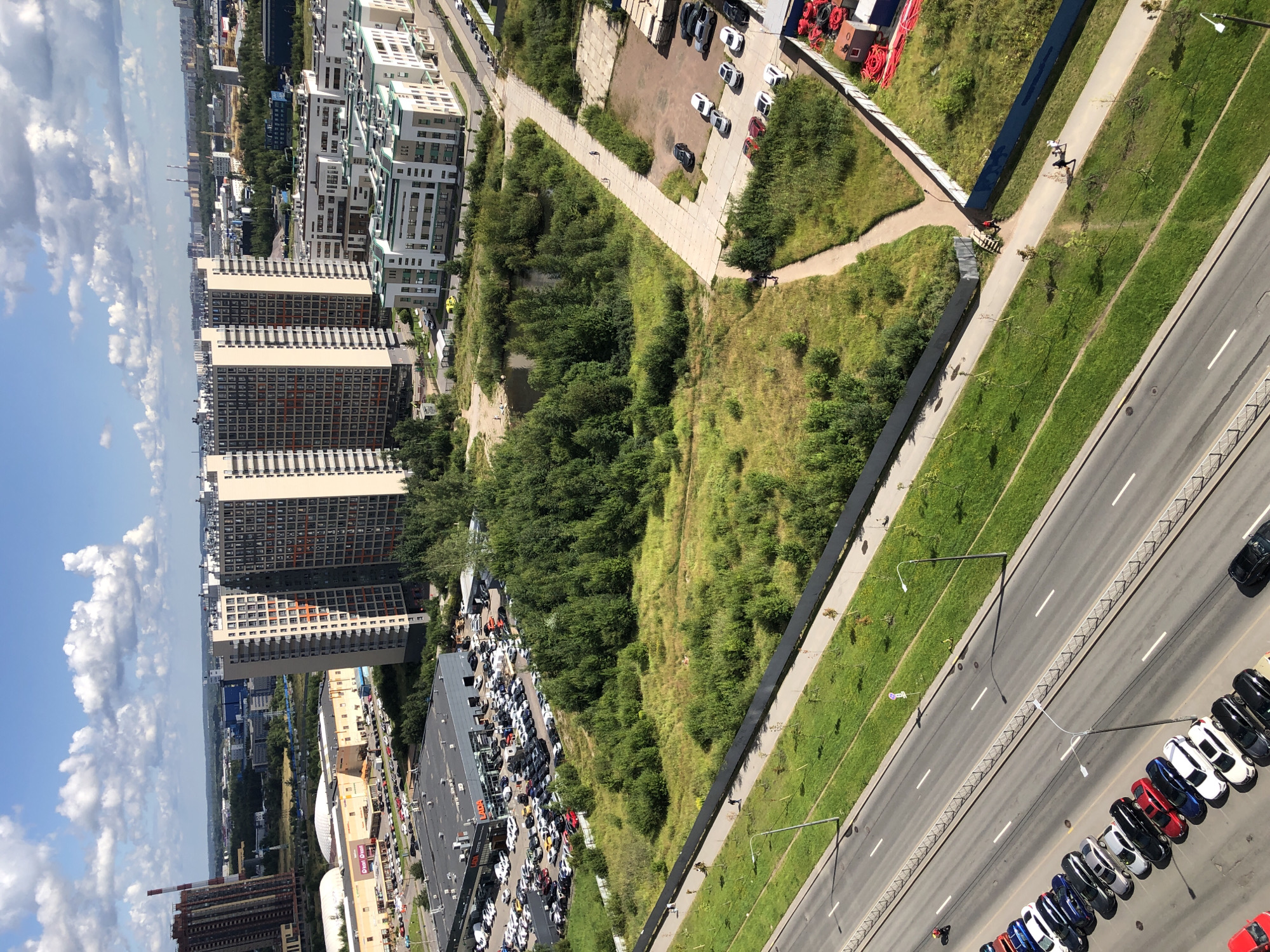 The image is an aerial view of a city scene on a sunny day with scattered clouds. In the foreground, a multi-lane road is visible with cars parked along one side and grass verges. A motorcycle is seen riding on the road. Further into the image, there's a large, undeveloped grassy area with a small pond, bordered by a fence. Beyond this area, several modern apartment buildings and a car dealership are situated. In the background, more urban structures and distant industrial or commercial buildings are apparent under a blue sky. Several individuals are walking on pathways near the grassy area and road.