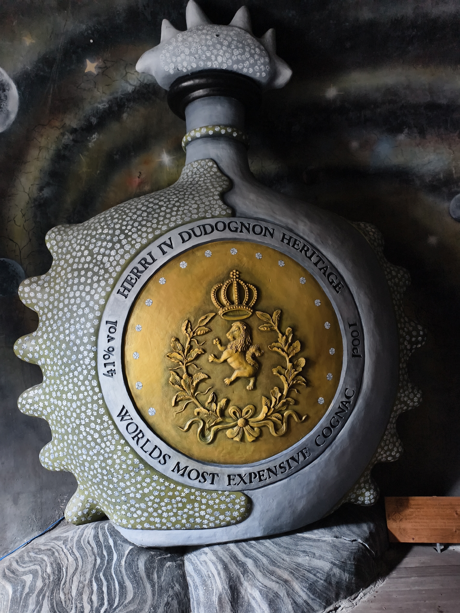 The image shows a large, ornate display resembling a bottle of Henri IV Dudognon Heritage Cognac, advertised as the "World's Most Expensive Cognac." The bottle is fabricated from a gray-white material covered in small white polka dots, with a large, gold-colored circular emblem in the center that features a crown, a lion within a laurel wreath, and the inscription "HERRI IV DUDOGNON HERITAGE" and "WORLDS MOST EXPENSIVE COGNAC." The bottle's details include "41% vol" and "100cl" markings. The entire display sits on a textured, gray-white base that mimics the appearance of stone or marble. The background features a dark mural with celestial elements like stars and a moon, suggesting a cosmic or otherworldly theme. A small wooden shelf or structure is visible to the right of the bottle. The location is Tay Nha Trang Ward, Vietnam.