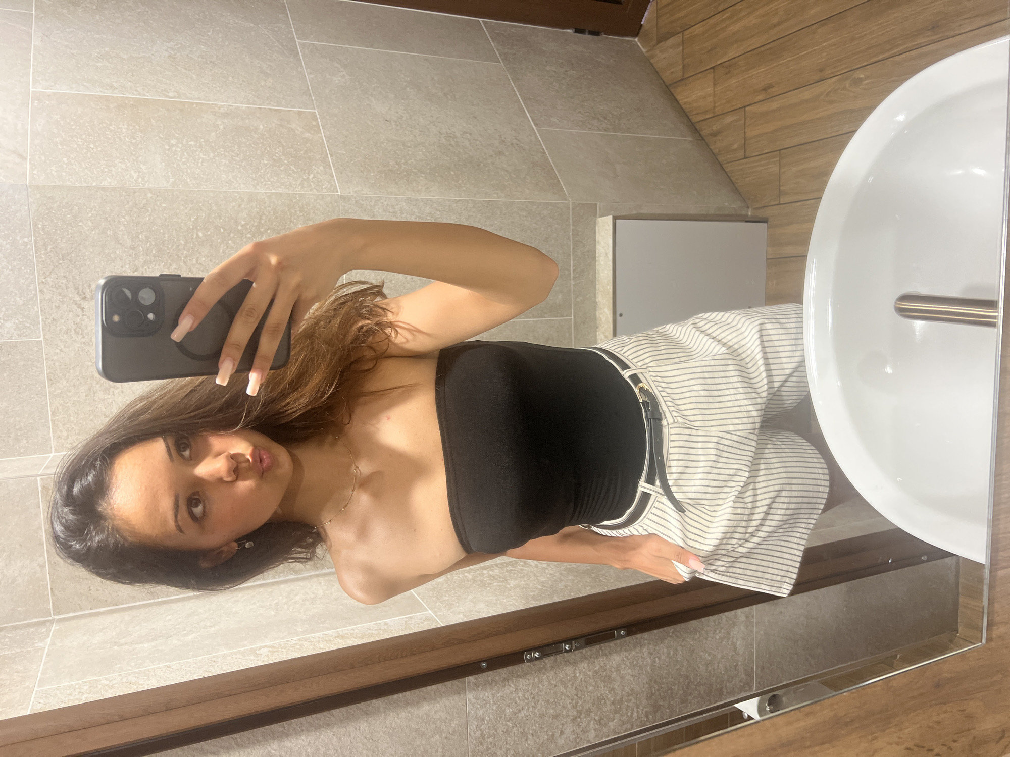 A young woman with long, brown hair is taking a mirror selfie in a bathroom. She is wearing a black strapless top and white and grey pinstripe shorts with a black belt. Her left hand is in her pocket. The bathroom has beige tiled walls and a wooden floor. A white sink is visible in the foreground, reflecting the image. The scene is indoors and appears to be in a residential setting.