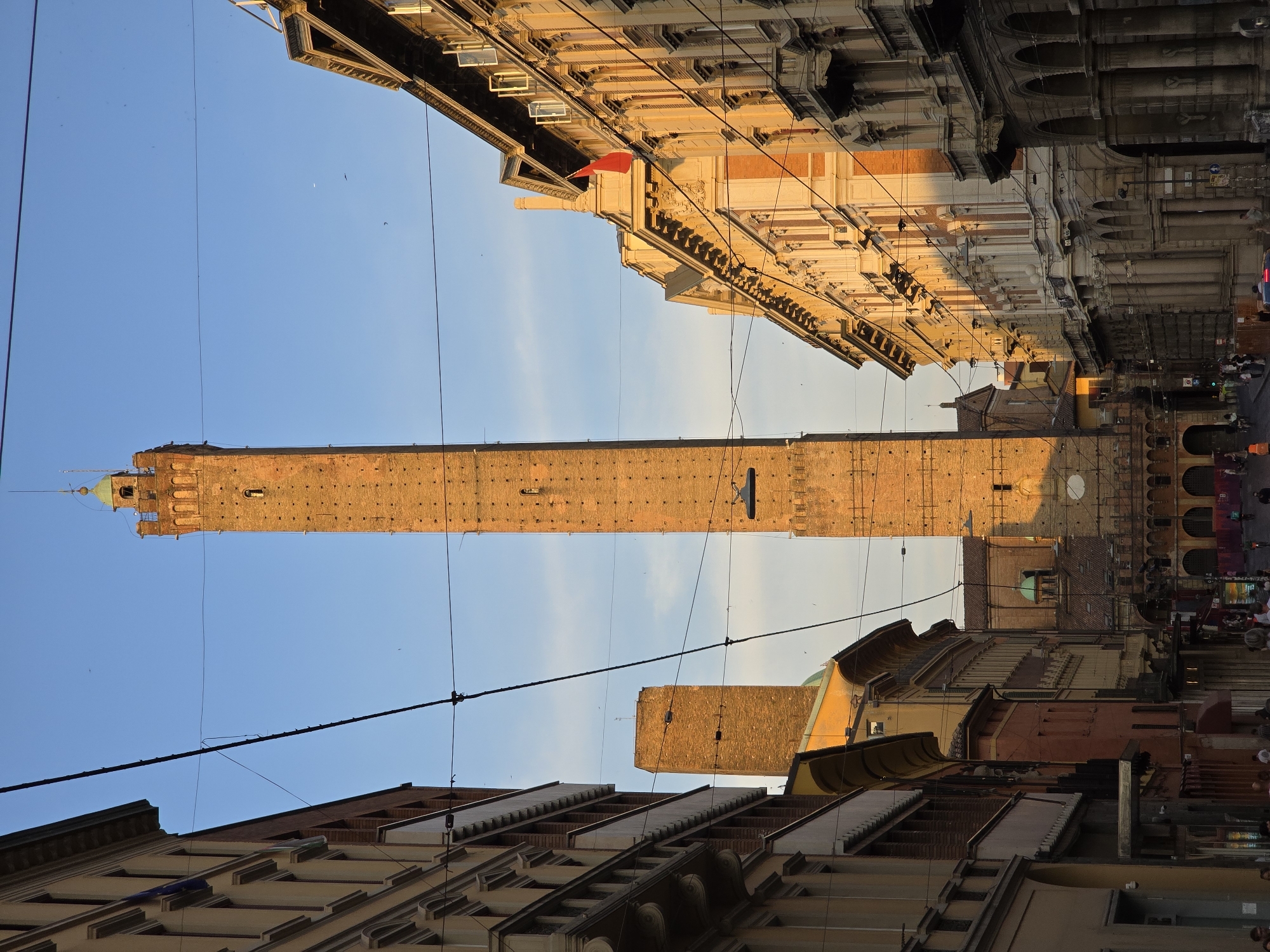 The image depicts a street scene in Bologna, Italy, dominated by the iconic Due Torri, specifically the Asinelli Tower, which stands tall and slender on the right side of the frame, bathed in the warm glow of the setting sun. Its brickwork is detailed with numerous small windows. To the left of the Asinelli Tower, the Garisenda Tower is visible, appearing shorter and leaning slightly. The towers are framed by the architecture of the city. On the left, a building with multiple stories and balconies lines the street, its facade displaying architectural details and windows. On the right, a grand building with ornate columns and arched walkways lines the opposite side of the street. The sky above is a clear blue with a few faint clouds. Several electrical wires crisscross the sky, creating a sense of urban infrastructure. In the foreground, the street is populated with people walking and some vehicles, indicating a lively urban environment. A red flag is visible on one of the buildings on the right. The overall atmosphere is one of historical grandeur and urban life under a beautiful sky.