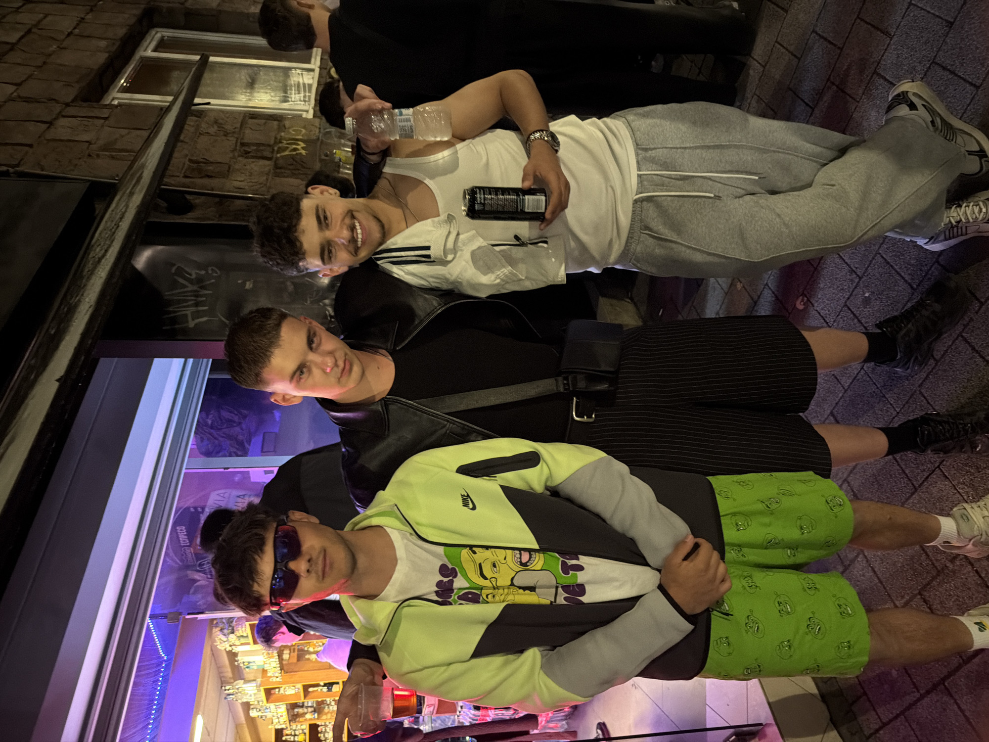 Three young men are posing for a picture outside what appears to be a bar or pub. The first man, closest to the camera, wears dark sunglasses, a lime green and grey Nike jacket over a white t-shirt with a graphic design, and green shorts with a cartoon-like print. The second man, in the middle, wears a black leather jacket over a black shirt and striped shorts. The third man, furthest from the camera, wears a white tank top and grey sweatpants. He holds a water bottle in one hand and a black can in the other. They are standing on a cobblestone street, and the background shows a building with brick walls and lit windows. The establishment behind them has purple lighting and shelves stocked with bottles. Based on the style of buildings and street, the location is likely in a city, specifically in Sofia, Bulgaria.