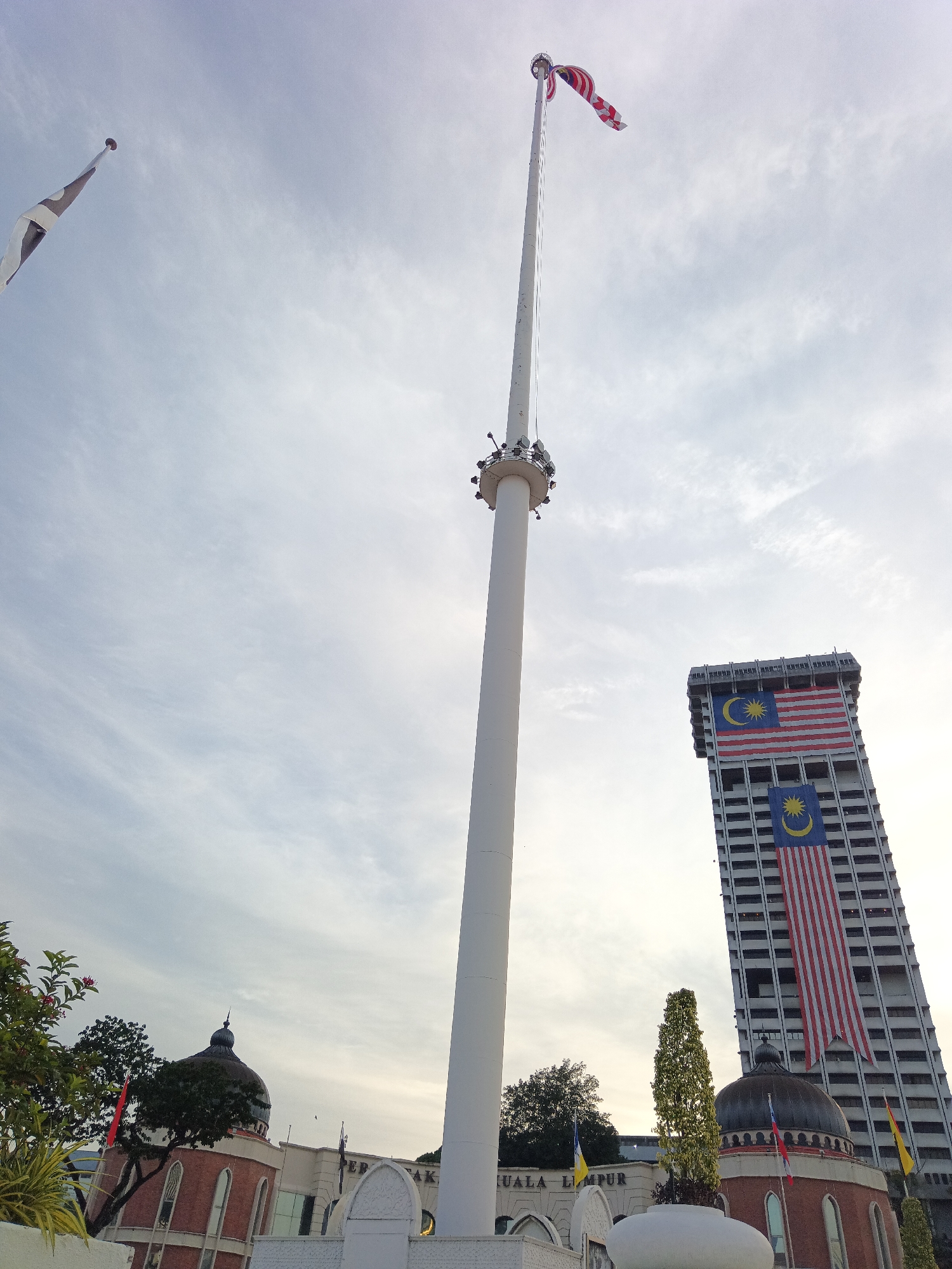 The image shows the Merdeka Square (Independence Square) in Kuala Lumpur, Malaysia. Dominating the scene is a very tall flagpole with a Malaysian flag flying at its apex, slightly to the right of the center. To the right of the flagpole, a tall, modern building is draped with two large Malaysian flags, one above the other. In the foreground and to the left, a domed building with red brick accents is visible, with trees and foliage in front of it. The sky is overcast with thin clouds. The overall impression is of a patriotic and monumental setting.