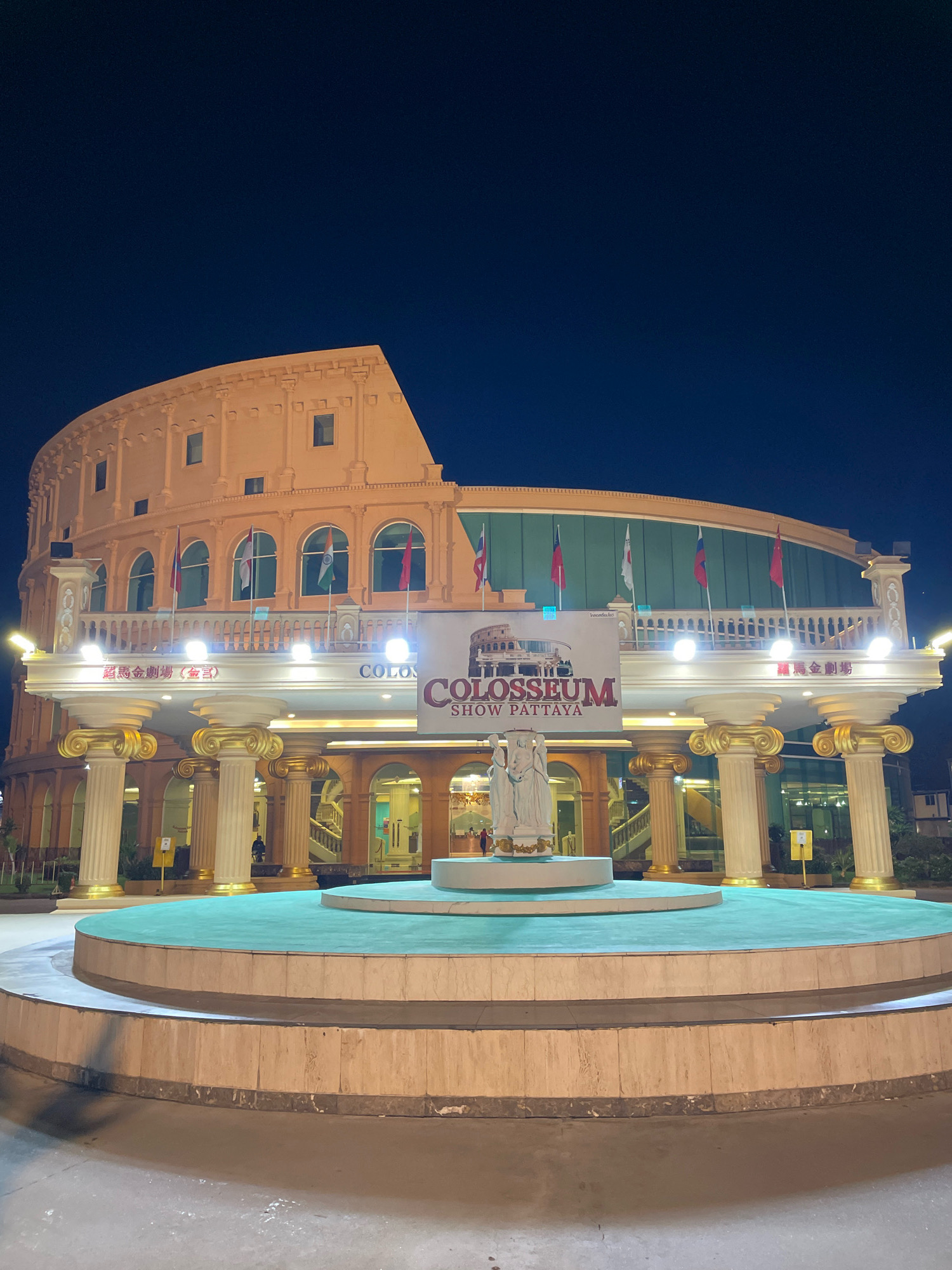 The image shows the exterior of the Colosseum Show Pattaya, a cabaret and performance venue located in Pattaya City, Thailand. The building is designed to resemble the Roman Colosseum and is illuminated at night. In the foreground, there is a multi-tiered circular fountain with a turquoise water surface. In the center of the fountain is a white sculpture of a group of figures. Behind the fountain, a grand entrance to the venue is visible, supported by large columns with ornate golden capitals. Above the entrance, a large banner displays "COLOSSEUM SHOW PATTAYA" along with an image of the Roman Colosseum. Several flags are mounted on a railing above the entrance. The sky is dark blue, indicating nighttime. There are a few people visible near the entrance and within the building's illuminated interior. The overall scene suggests a nighttime tourist attraction.