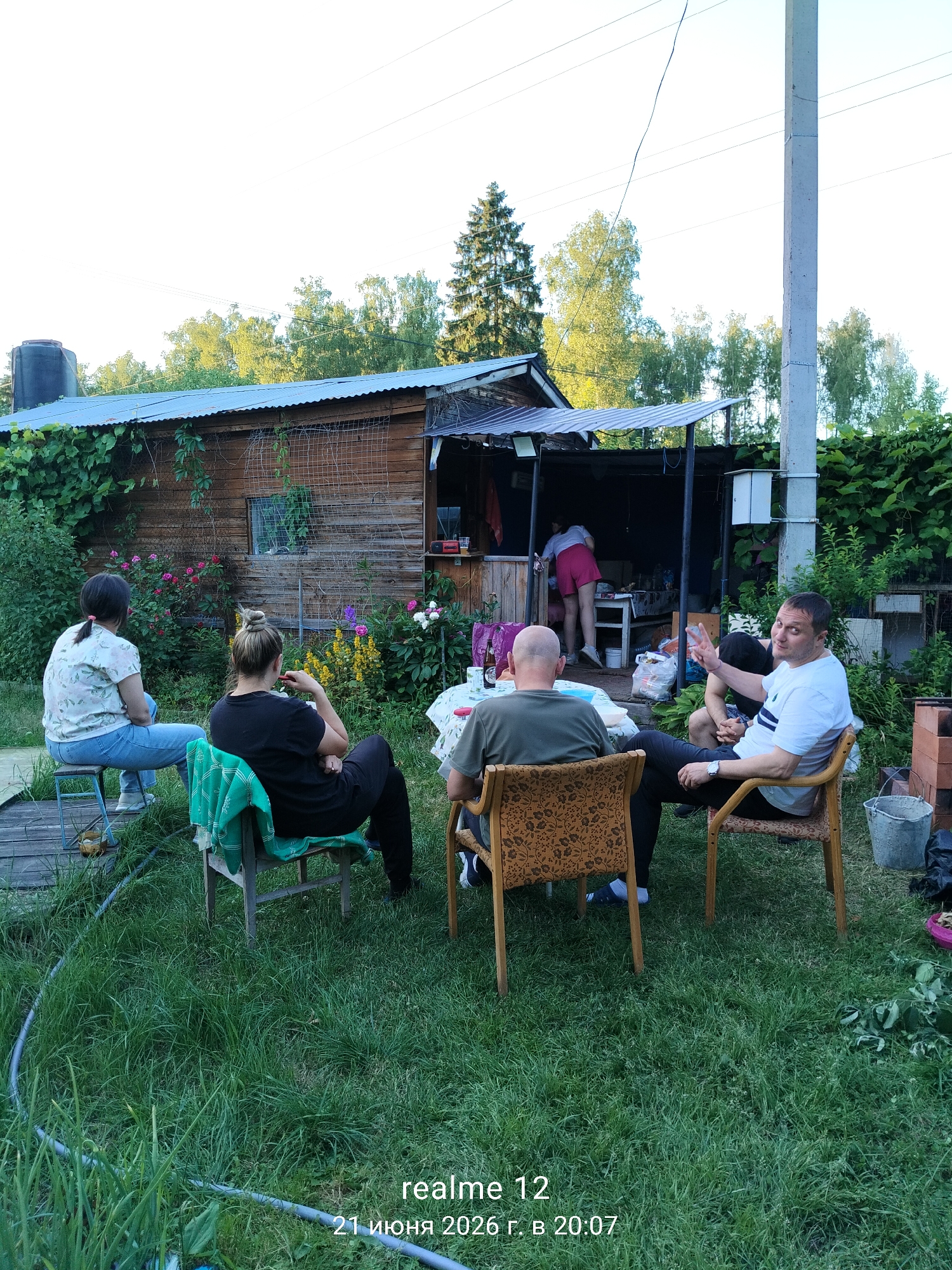 The image depicts a backyard gathering in the city of Космос-2 Снт, Russia. Several people are seated on chairs around a table outdoors. In the background, a wooden shed with a corrugated metal roof is visible, surrounded by lush greenery and flowering plants. One person is standing inside a covered outdoor area attached to the shed. The atmosphere appears casual and relaxed, suggesting a social gathering.