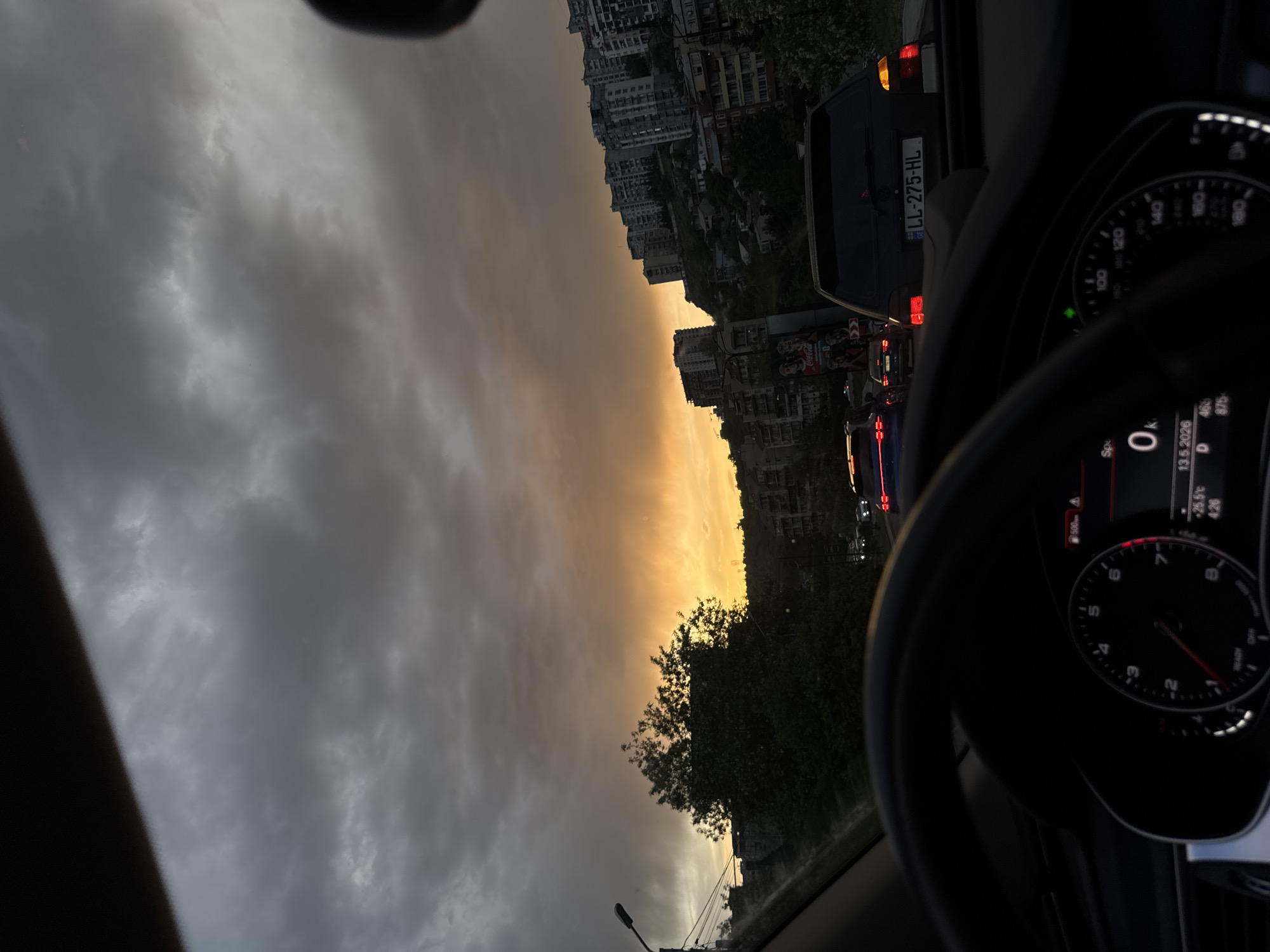 The image, taken from inside a car, shows a street scene in Tbilisi, Georgia at sunset on 13.5.2026. The car is stationary, as indicated by the speedometer reading "0 km." The dashboard display shows the temperature as 25.5°C and the time as 4:26. Other cars are visible in the traffic ahead, including one with the license plate "LL-275-HL." The cityscape is visible in the background, with buildings silhouetted against the orange sky. The sky is partially obscured by heavy, grey clouds. A Coca-Cola advertisement is visible beside the car.