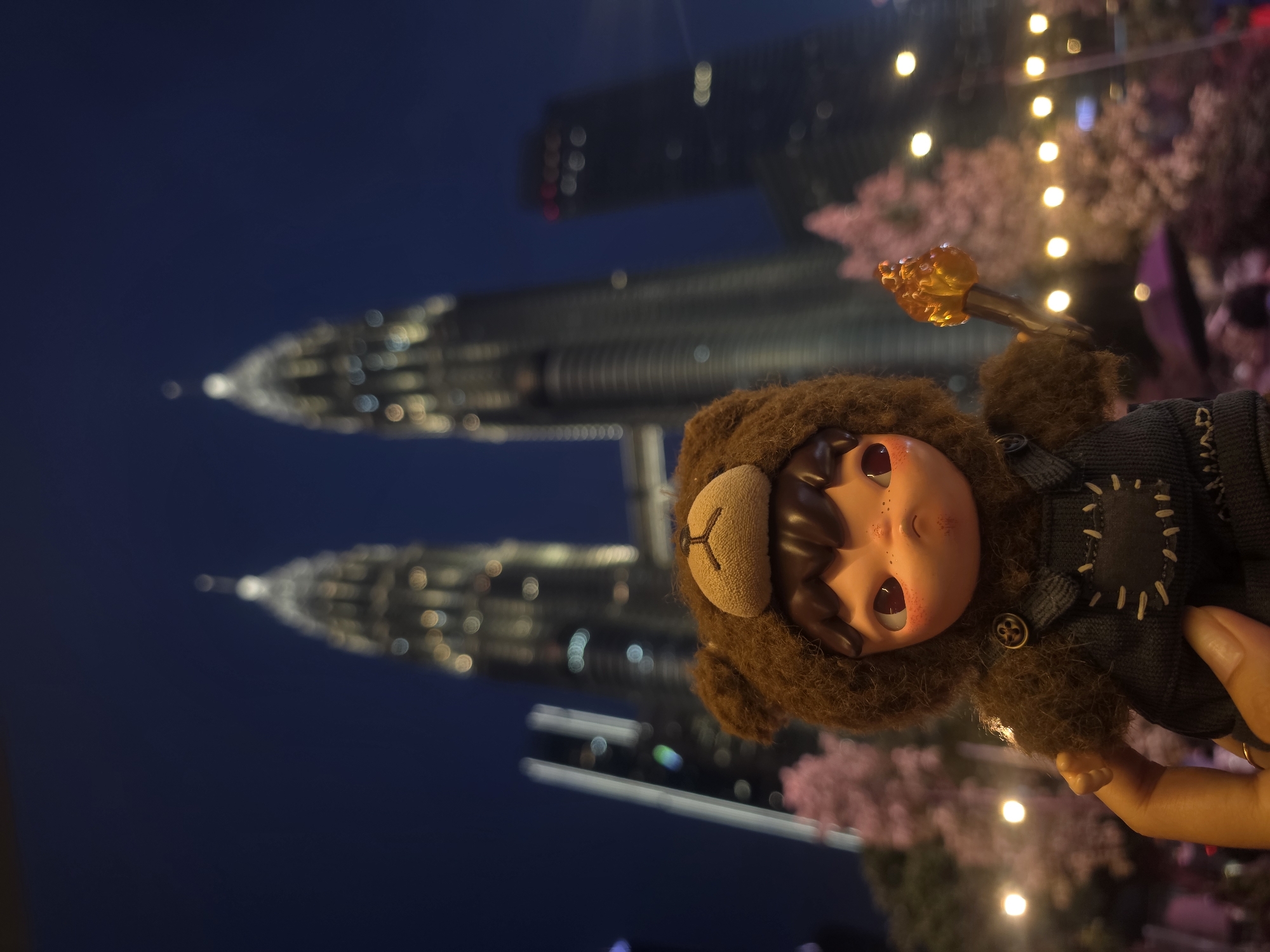 In Kuala Lumpur, Malaysia, at night, a doll dressed in a bear costume and carrying a torch is held in a hand. In the blurry background are the illuminated Petronas Twin Towers and blurred lights suggestive of trees or decorations. The sky is dark blue.