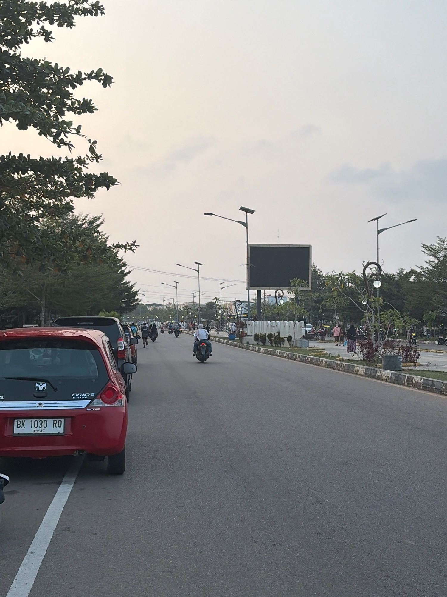 The image depicts a street scene in Tanjung Pinang, Indonesia, possibly during dusk or early morning, given the soft lighting and overcast sky. In the foreground on the left, a red car with the license plate "BK 1030 RO" is parked. The road stretches into the distance, lined with trees and streetlights. Several motorcycles are visible on the road, with riders in motion. In the mid-ground, a large, dark rectangular screen is mounted on a pole, likely a digital billboard. Pedestrians can be seen walking along the sidewalk and across the street. The environment appears to be an urban or suburban setting with well-maintained infrastructure, including a paved road, sidewalks, and landscaping. The overall atmosphere is one of moderate activity and a relaxed urban environment.