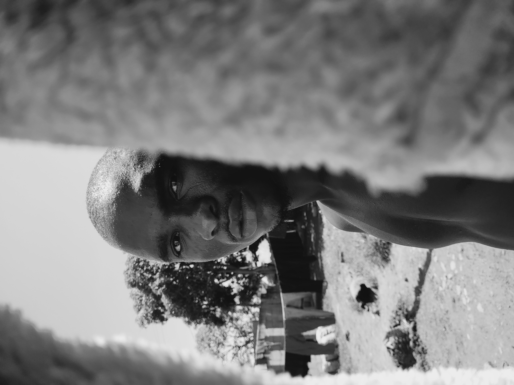 This black and white portrait captures a Black man in what appears to be an outdoor setting in Bukuru, Nigeria. The man, likely in his 20s or 30s, is positioned in the mid-ground, looking directly into the camera with a neutral and somewhat intense gaze. His head is shaven or has very short hair, and strong highlights on his forehead and shoulders suggest bright, direct daylight. He is bare-chested, with the curve of his upper body visible.
The background is softly blurred but reveals natural elements like trees with sparse leaves on the left and center, and what seems to be unpaved ground or simple structures on the right, indicative of a rural or semi-urban environment. The sky, visible in the upper left, is bright, confirming it's daytime.
A heavily blurred, textured object, resembling a fuzzy blanket or cloth, occupies the immediate foreground and background (top and bottom when viewed upright), framing the subject and adding a layer of depth. No specific activity is taking place other than the man posing for the photograph. There is no visible text in the image. The overall impression is one of directness and introspection.