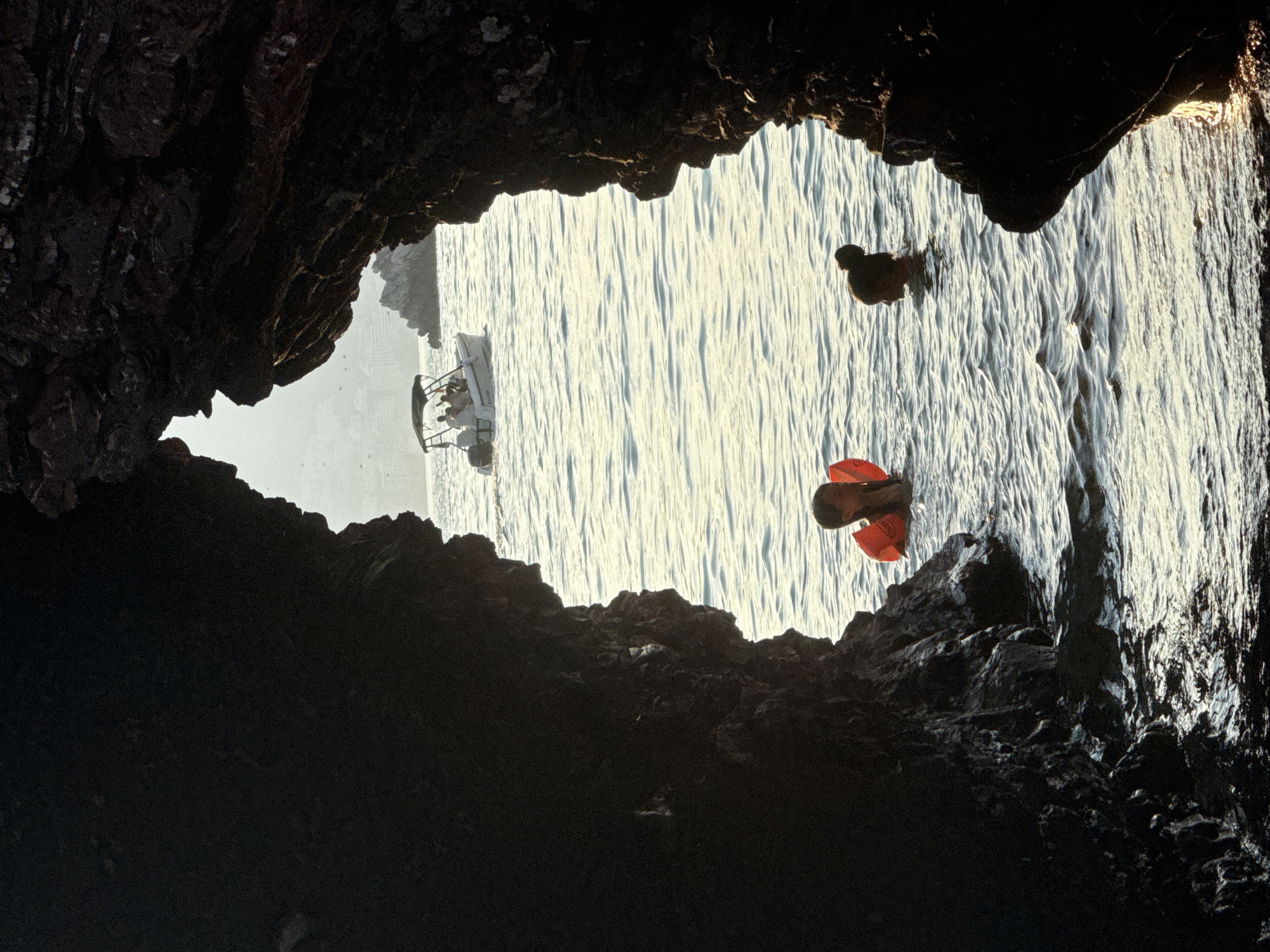 The image is a vertical, low-angle shot taken from within a dark cave or overhang, looking out onto a bright, rippling body of water. The foreground is dominated by the rough, dark texture of the rock formation, which frames the scene like a natural window.
In the water, a white boat with several people on board is visible in the distance, heading towards the right side of the frame. Closer to the foreground, two subjects are in the water. One appears to be a person with dark hair, possibly swimming or treading water, their upper body submerged. The other, closer to the bottom of the frame, is a person wearing an orange flotation device, their legs and the device visible above the water. The water itself is illuminated by bright sunlight, creating a textured surface with highlights and shadows.
The scene suggests a recreational activity, likely swimming or boating in the sea. The bright water and the presence of people in swimwear indicate it is daytime, probably late morning or early afternoon, given the brightness of the light. The backdrop shows land or cliffs in the distance, suggesting a coastal location. The overall mood is serene, with the natural cave framing a peaceful scene of leisure on the water. No visible text is present in the image. The location context is Budva, Montenegro.