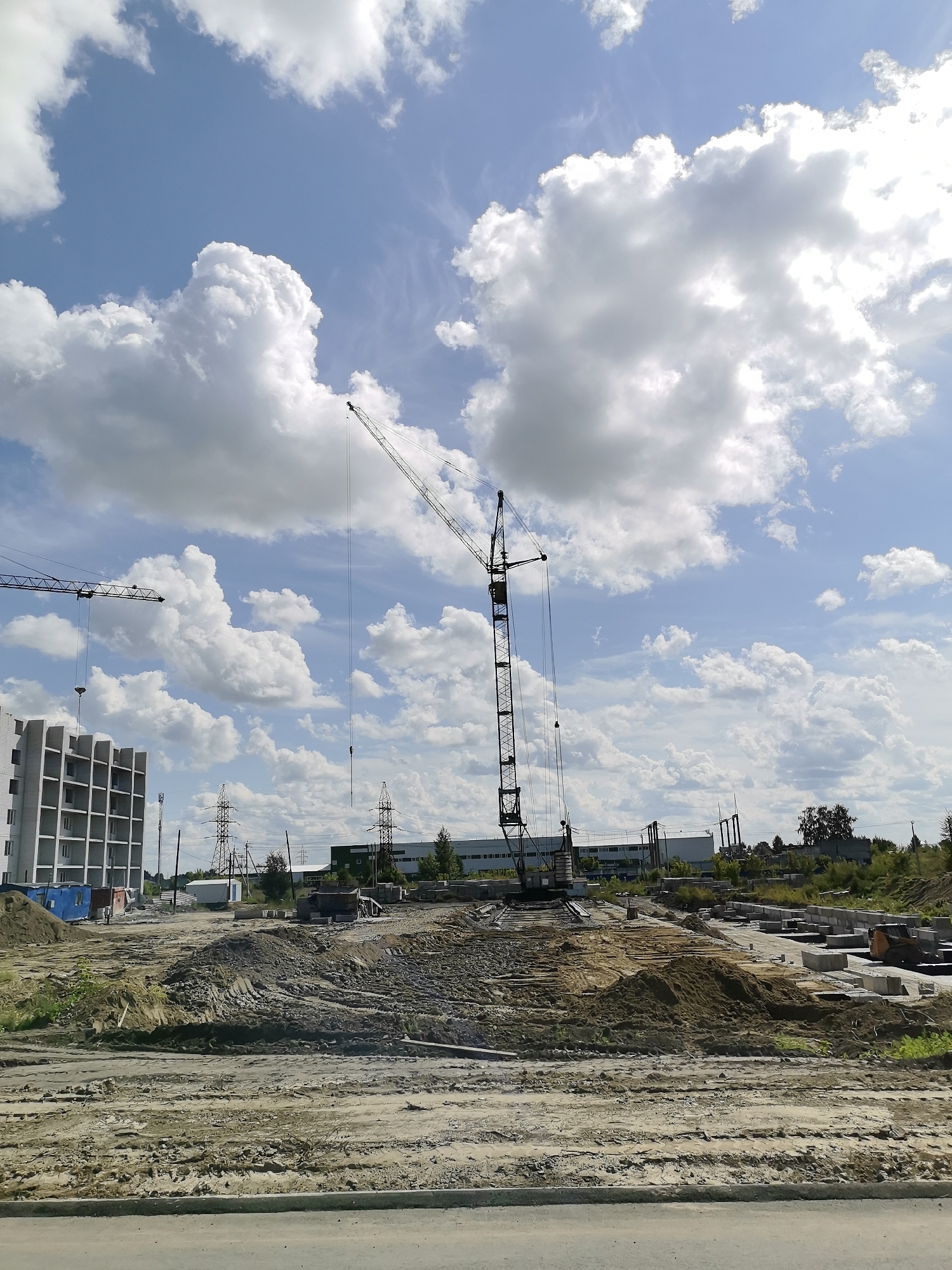 The image depicts an outdoor construction site on a cloudy day. In the foreground, a rough dirt path or road is visible. Behind it, a construction area is characterized by mounds of dirt and sand, as well as stacks of concrete blocks. Several industrial buildings and power lines are situated further back. Two large construction cranes dominate the scene; one is a tall, slender tower crane in the center of the frame, and another, a boom crane, is visible on the left. A multi-story building, partially constructed and appearing to be made of white bricks, stands to the left of the tower crane. The sky is a bright blue, filled with large, fluffy white clouds. The overall impression is one of ongoing construction in an industrial or semi-rural setting, under a dynamic sky. This scene is located in Novoaltaysk, Russia.