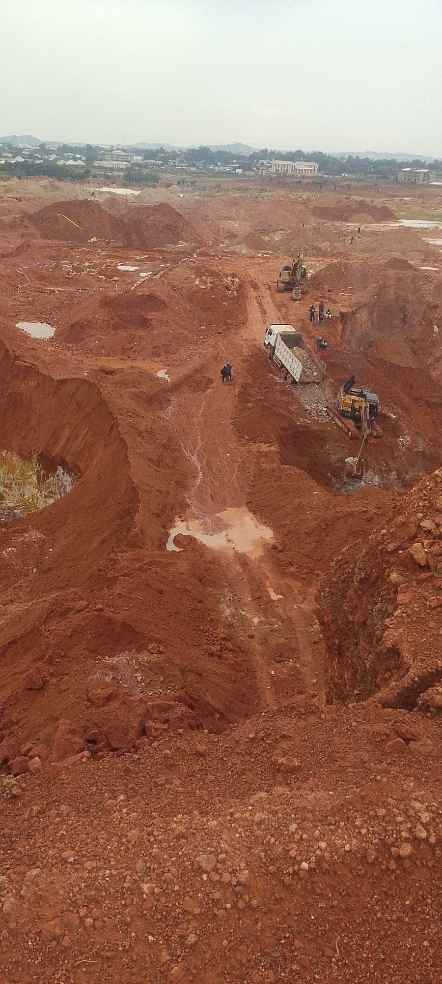 The image depicts an aerial view of a large, excavated construction site with reddish-brown earth. In the background, a distant city or town is visible with buildings and trees. The foreground is dominated by the excavated landscape, characterized by steep slopes, mounds of dirt, and some puddles of water. Several pieces of heavy construction equipment are present, including a large excavator with its arm extended and a dump truck being loaded. There are also a few individuals visible on the site, appearing to be workers, some of whom are near the vehicles. The overall scene suggests ongoing excavation or construction work in an area that was once a natural landscape. The city of Reifild-Ist, Nigeria, is a town in the Northern Region of Nigeria, known for its large agricultural base and growing infrastructure. The construction site appears to be on the outskirts of the developed area, with the natural landscape giving way to large-scale earthmoving operations.