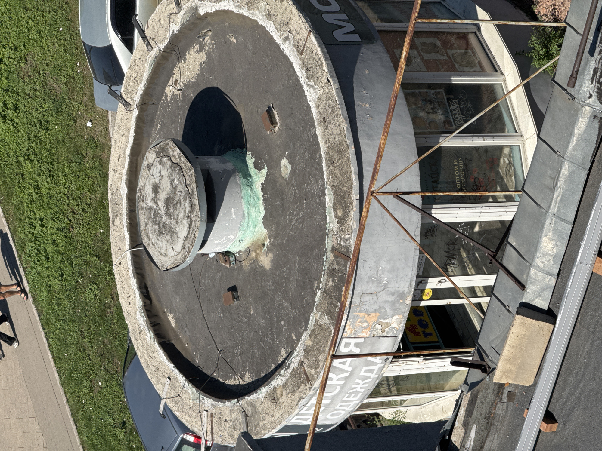 The image is an aerial view of a round, flat-roofed building with a vent pipe in the center, located in Krasnoyarsk, Russia. The building appears to be a shop or business, with large windows displaying signs and advertisements. One sign reads "ДЕТСКАЯ ОДЕЖДА" (children's clothing), and another has text that includes "ОПТОМ И В РОЗНИЦУ" (wholesale and retail). There are cars parked near the building, one a light blue sedan and another a darker sedan. In the background, a paved walkway and a grassy area are visible, with two people walking on the walkway. The overall scene depicts an urban environment on a sunny day.